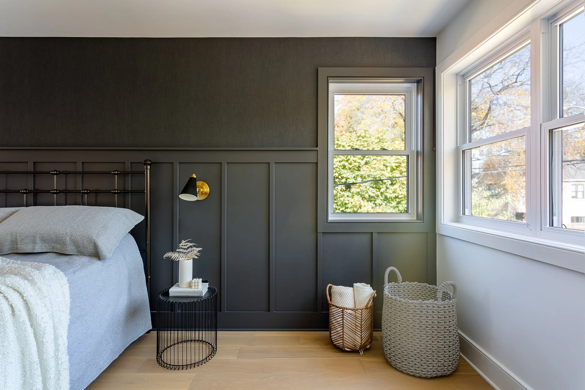 Bedroom with dark gray accent wall, bed with gray bedding, black and gold wall-mounted reading lamp, small black side table with white decor, wicker basket and woven basket near window, view of autumn trees outside