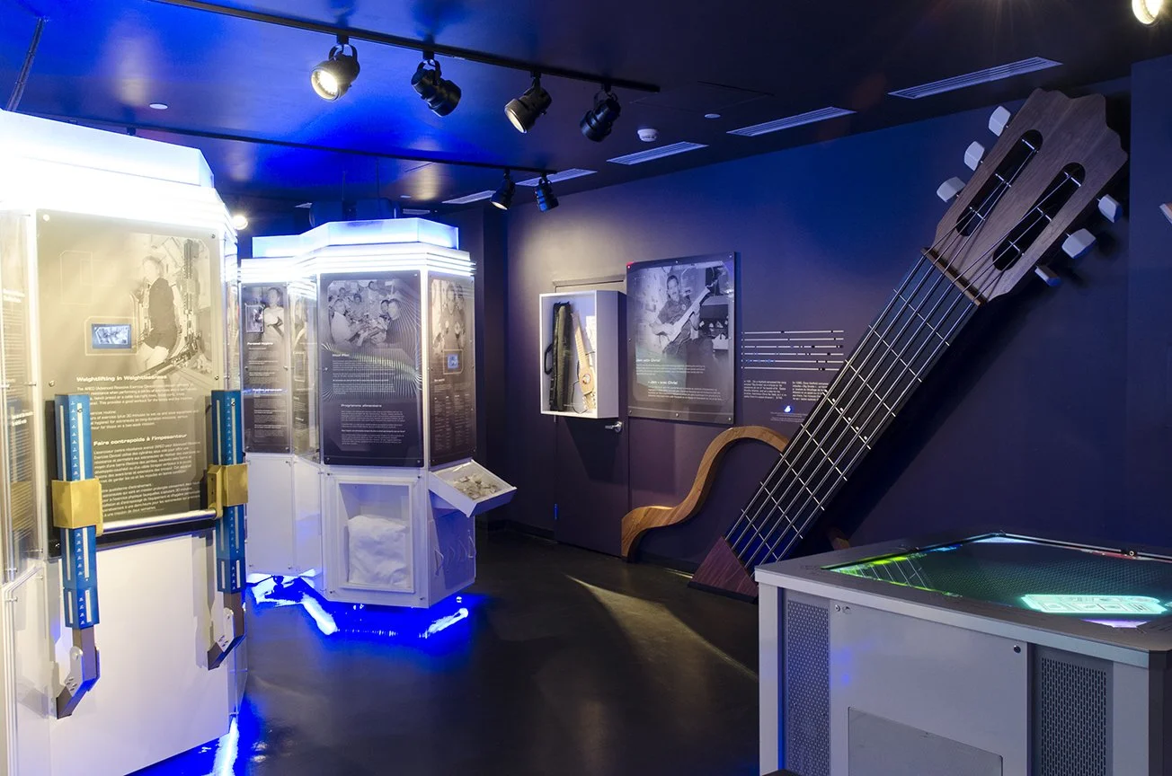 Exhibit in a museum with illuminated display cases, a giant guitar sculpture on the wall, and a pinball machine with colorful lights.
