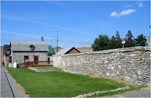 A grassy area next to a stone wall in a residential neighborhood with houses in the background under a clear blue sky.