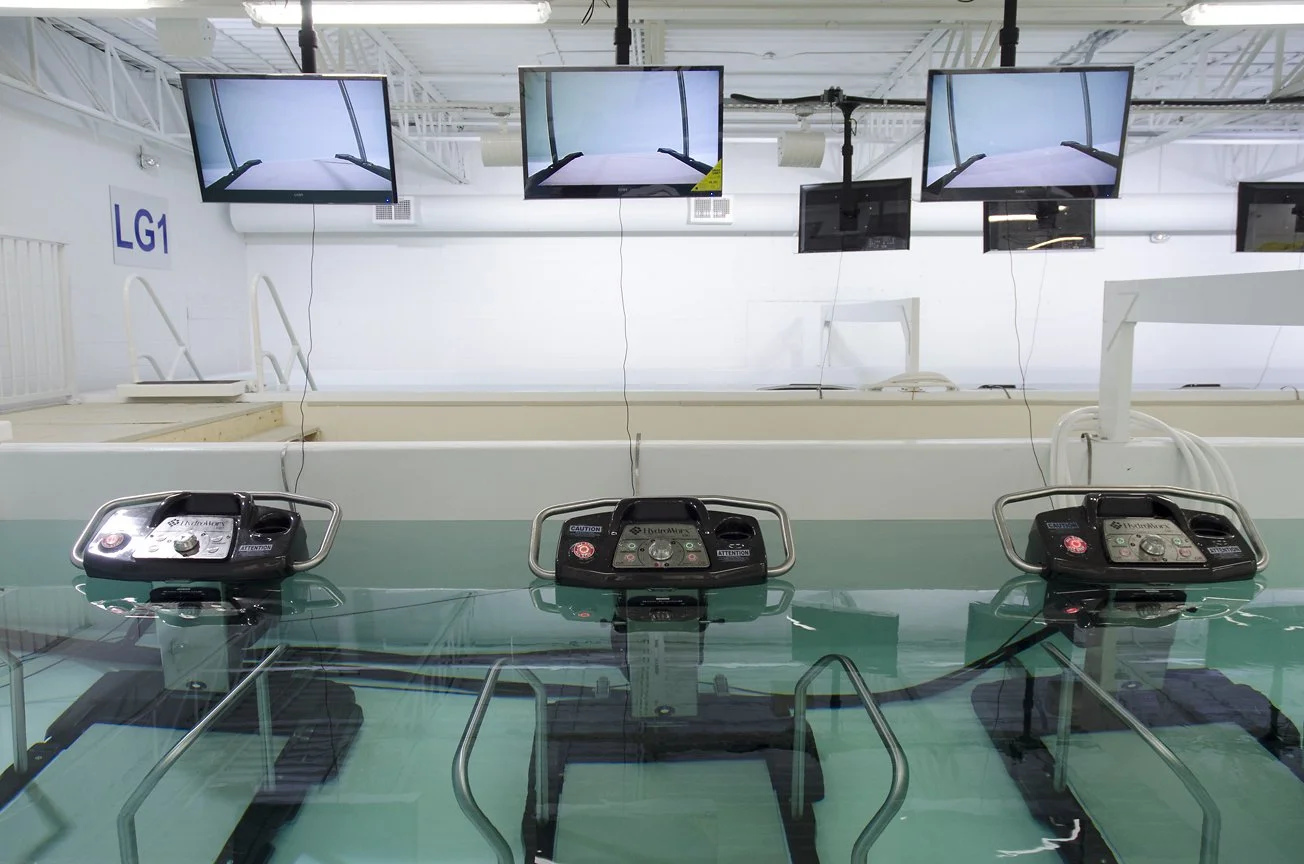 Three medical devices are submerged in a tank of water in a controlled environment, with multiple monitors overhead displaying different angles of the setup.