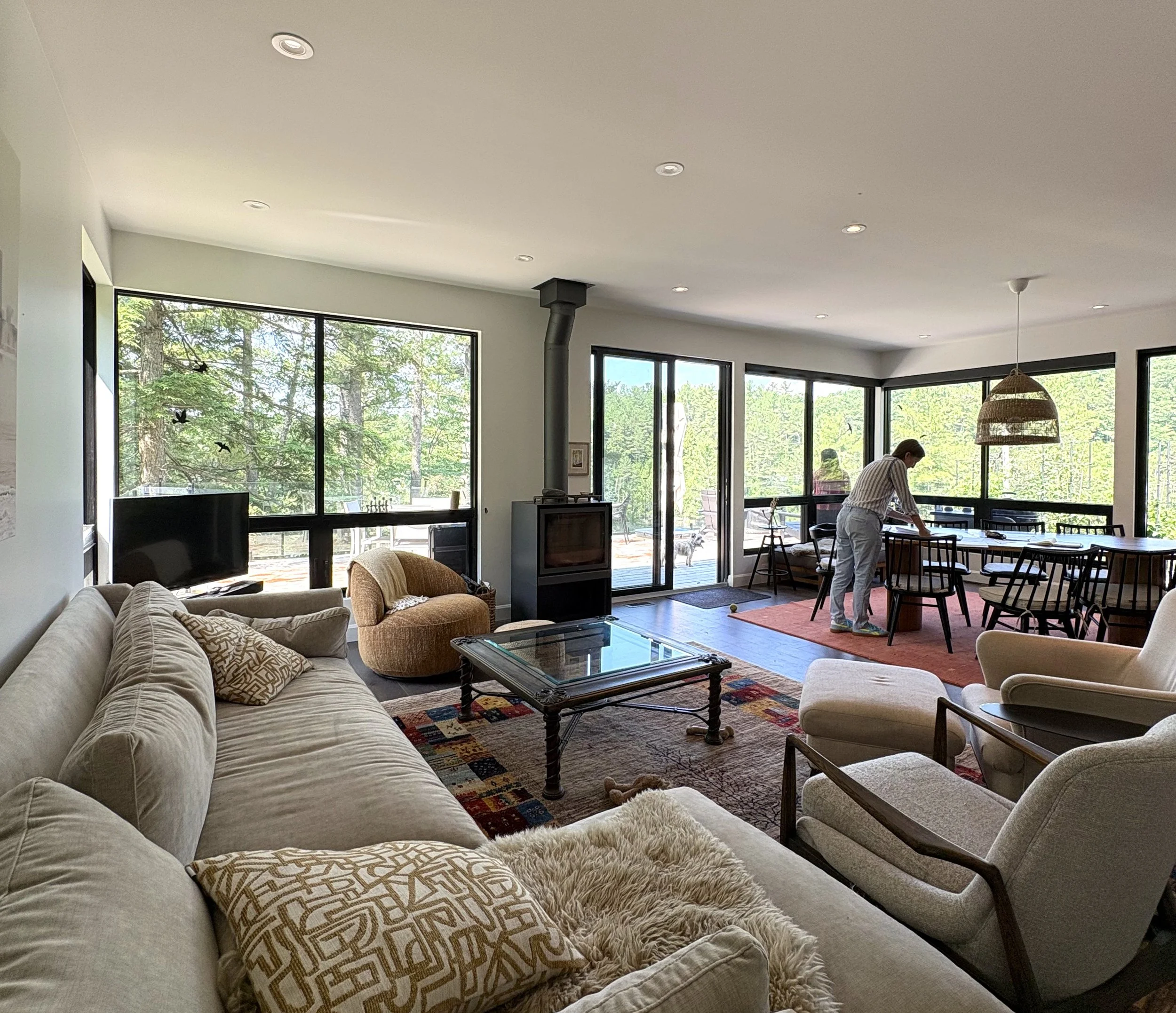 Living room with large windows overlooking a wooded outdoor area, containing a beige sofa with patterned pillows, an orange armchair, a glass-top coffee table, and a dining area where a person is setting the table. There's also a wood stove and a television.