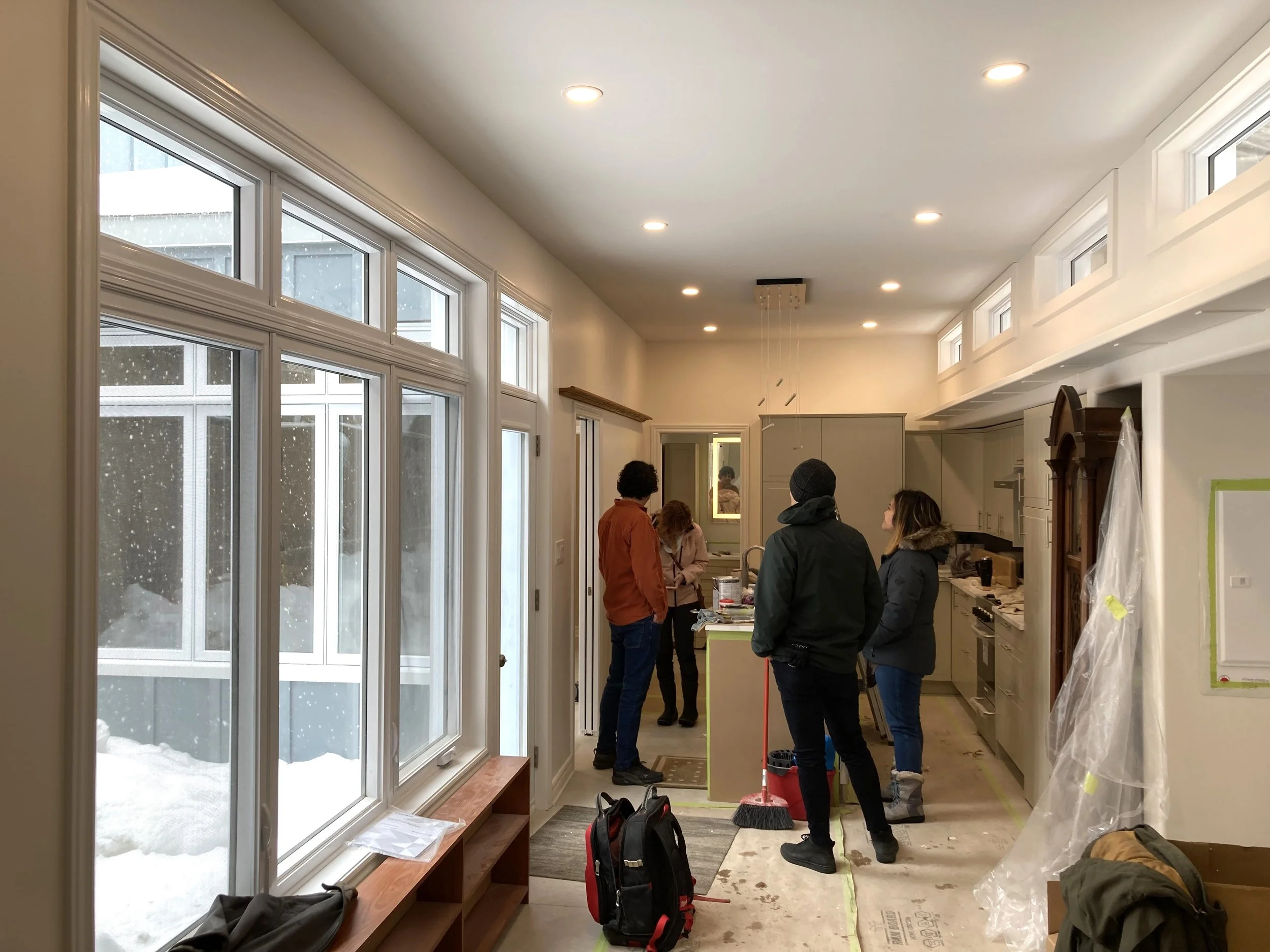 A group of people inside a kitchen under renovation, with large windows showing snow outside, and construction materials and tools around.