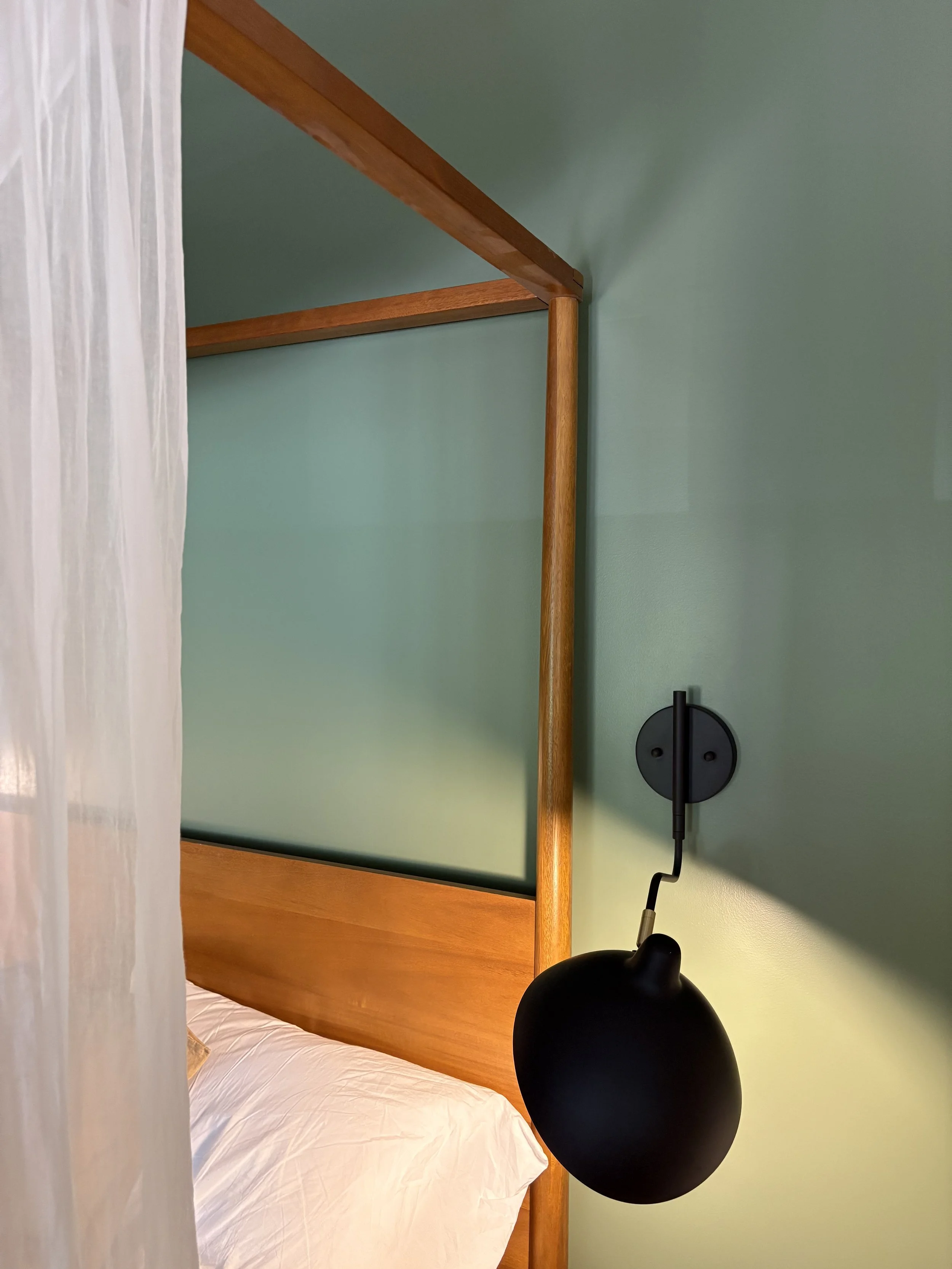 Close-up of a black wall-mounted reading lamp beside a wooden bed with a white pillow and bed sheet, with a sheer curtain on the left and a green wall in the background.