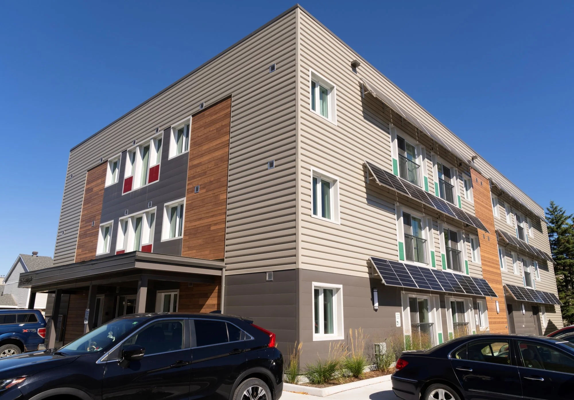 Modern multi-story apartment building with solar panels on the windows and contemporary exterior design, including wood and metal siding. Several cars are parked in front.