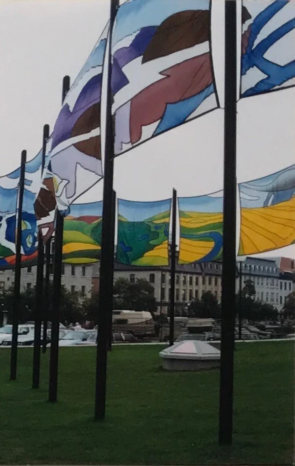 Colorful fabric banners hanging from tall poles in an outdoor park, with buildings and trees in the background. NCC banner competition winner Jane Thompson, 1988 Ottawa Ontario Canada.