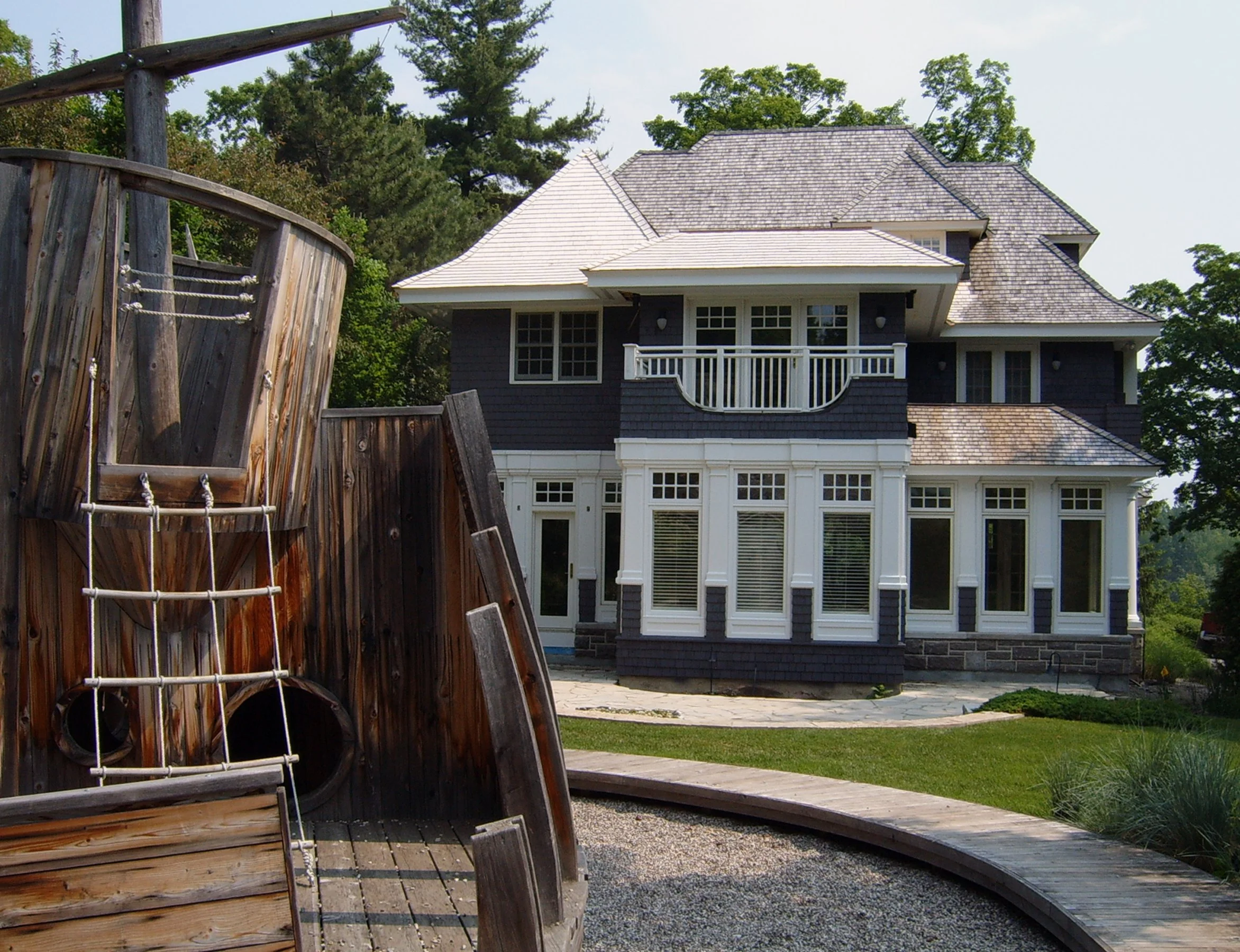 Front view of a large house with multiple windows, a small balcony, and a sloped roof, partially obscured by a wooden playground structure in the foreground.