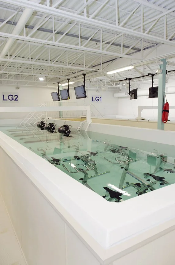 Indoor swimming pool with exercise machines submerged in water, viewing from the side. Overhead monitors and a high ceiling with exposed pipes.