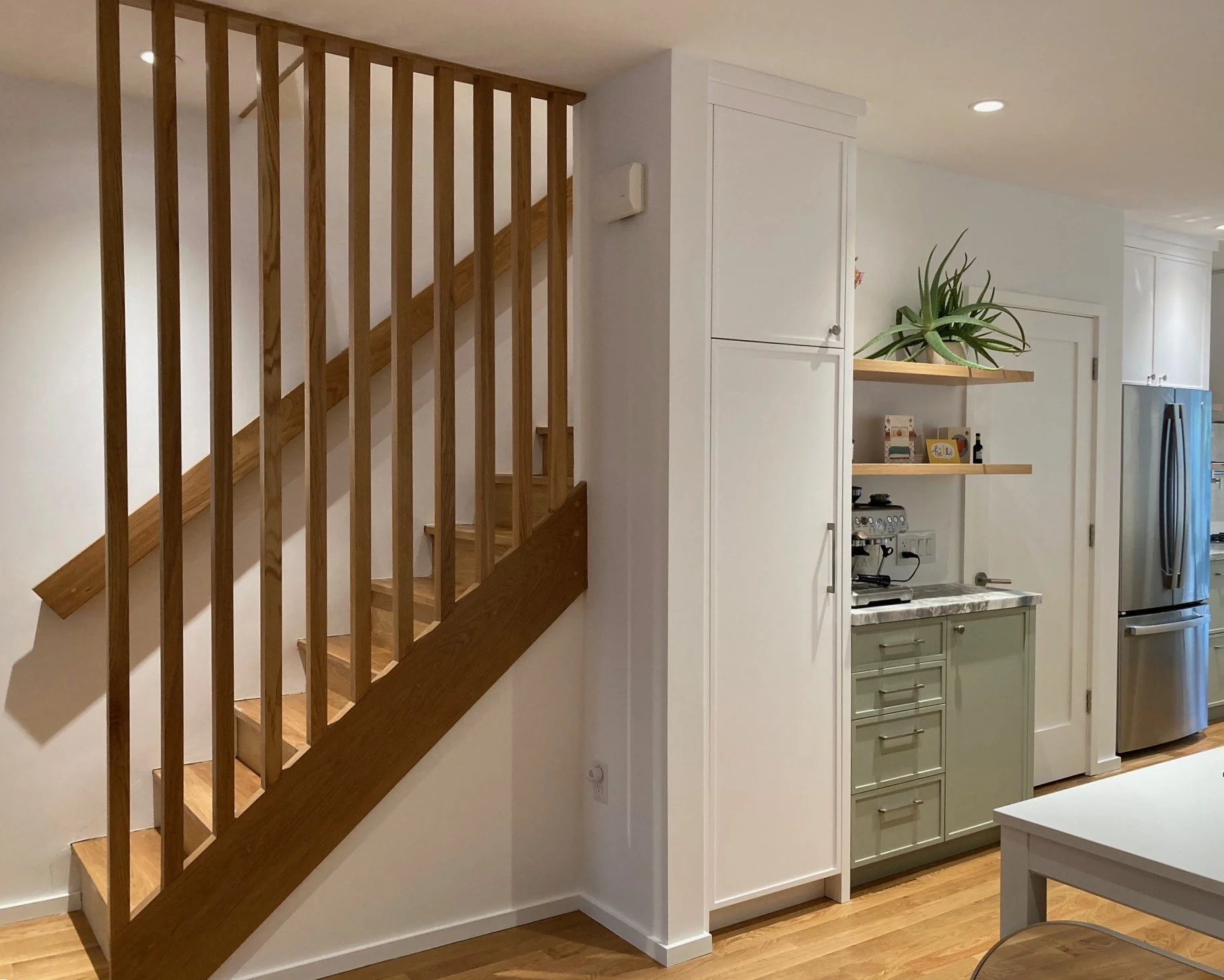 Interior of a home with a wooden staircase on the left, and a kitchen area on the right featuring white cabinets, open shelves with decorative items and a potted plant, and a stainless steel refrigerator.