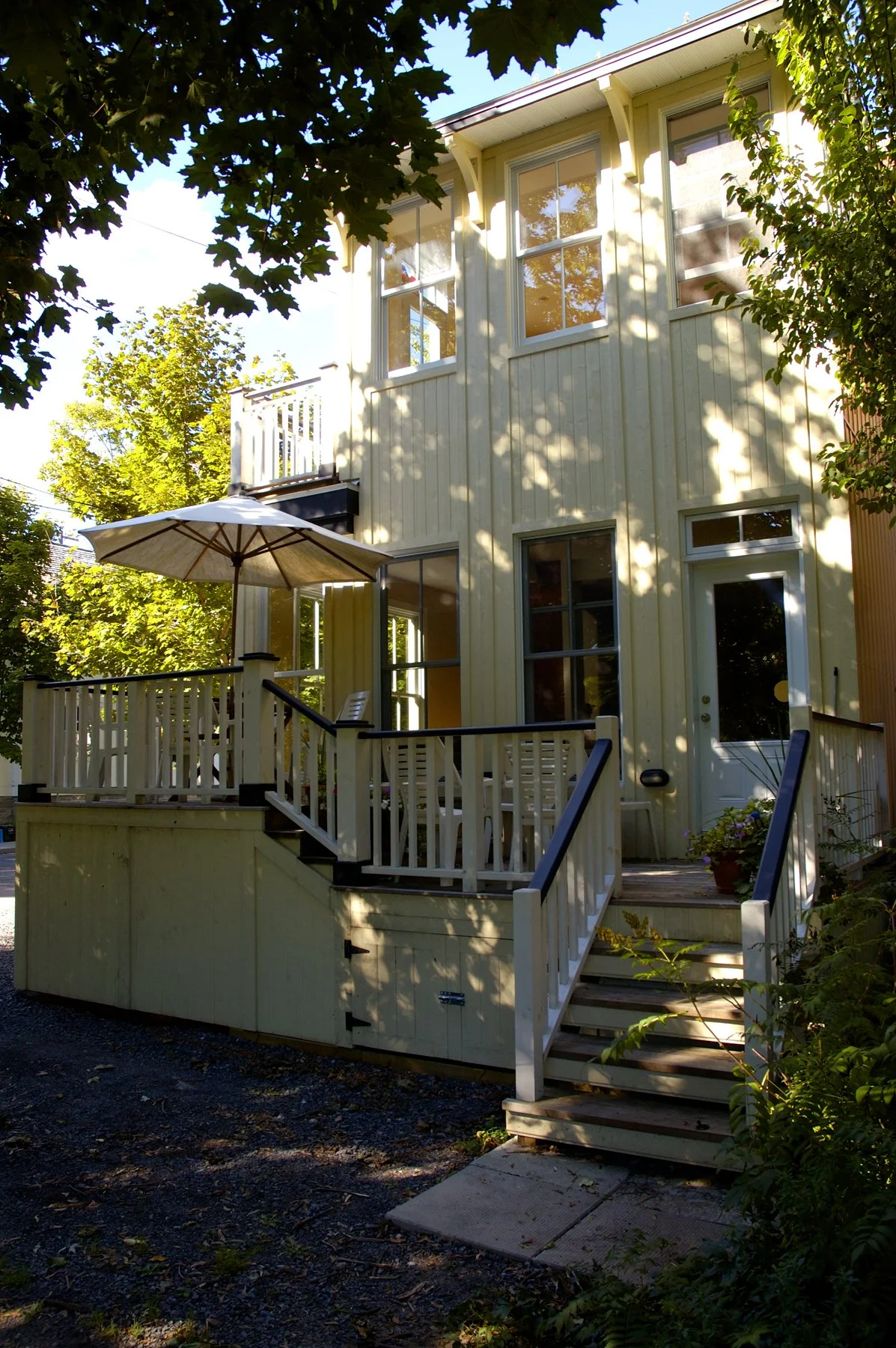 A two-story yellow house with multiple white-framed windows and a deck with white railing. There is an umbrella and outdoor furniture on the deck, surrounded by trees and shadows.