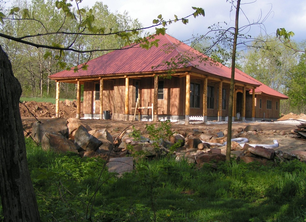 A house under construction with a red metal roof, wooden support beams, and unfinished exterior walls, surrounded by trees and rocks.