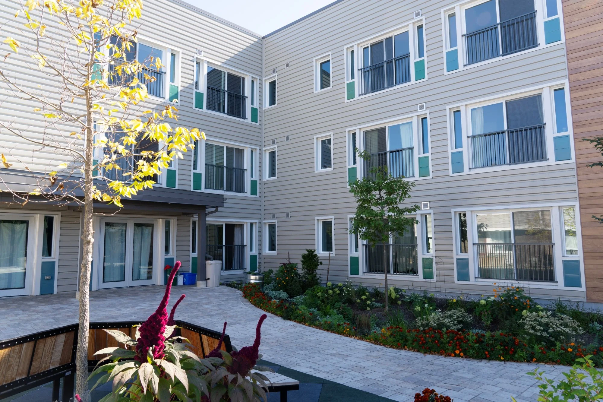 Open courtyard of a modern apartment complex with multiple windows and small balconies, landscaped with trees and colorful flowers, on a sunny day.