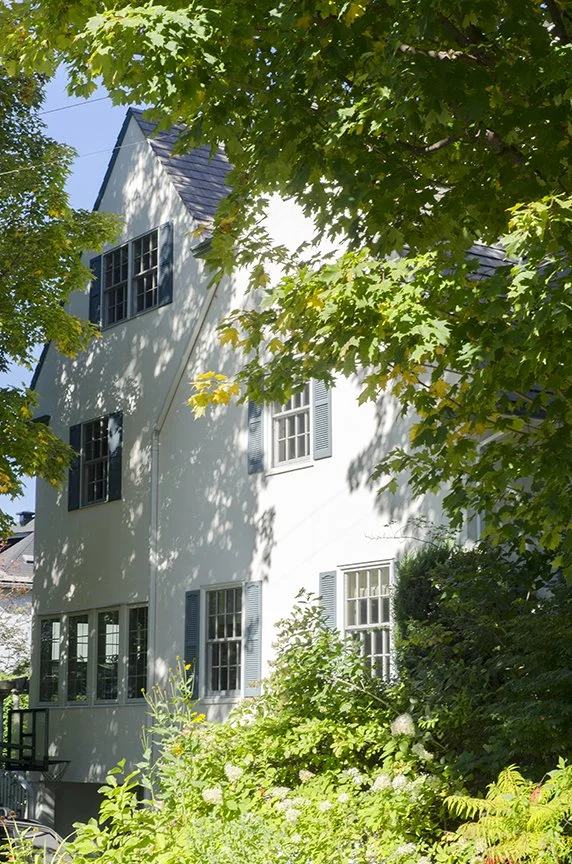 A white multi-story house with blue shutters, surrounded by green trees and bushes, with sunlight filtering through the leaves.