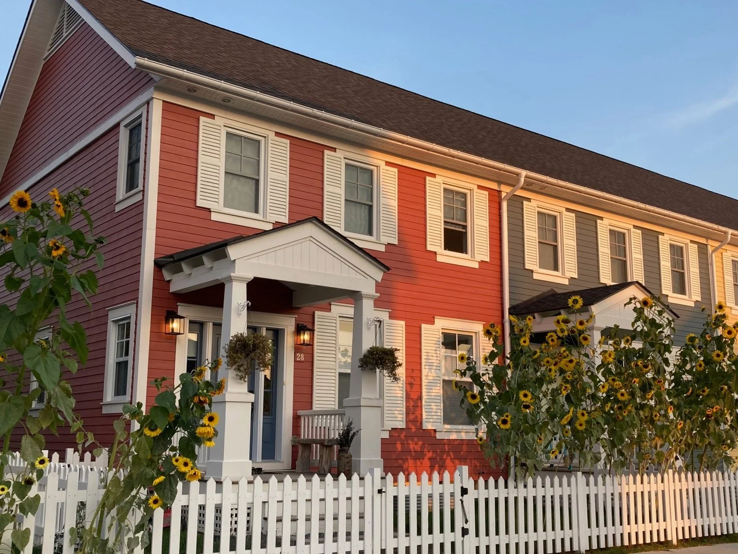 A two-story house with pink and blue exterior siding, white shutters, and a white picket fence, with sunflowers in front and the sky in the background.