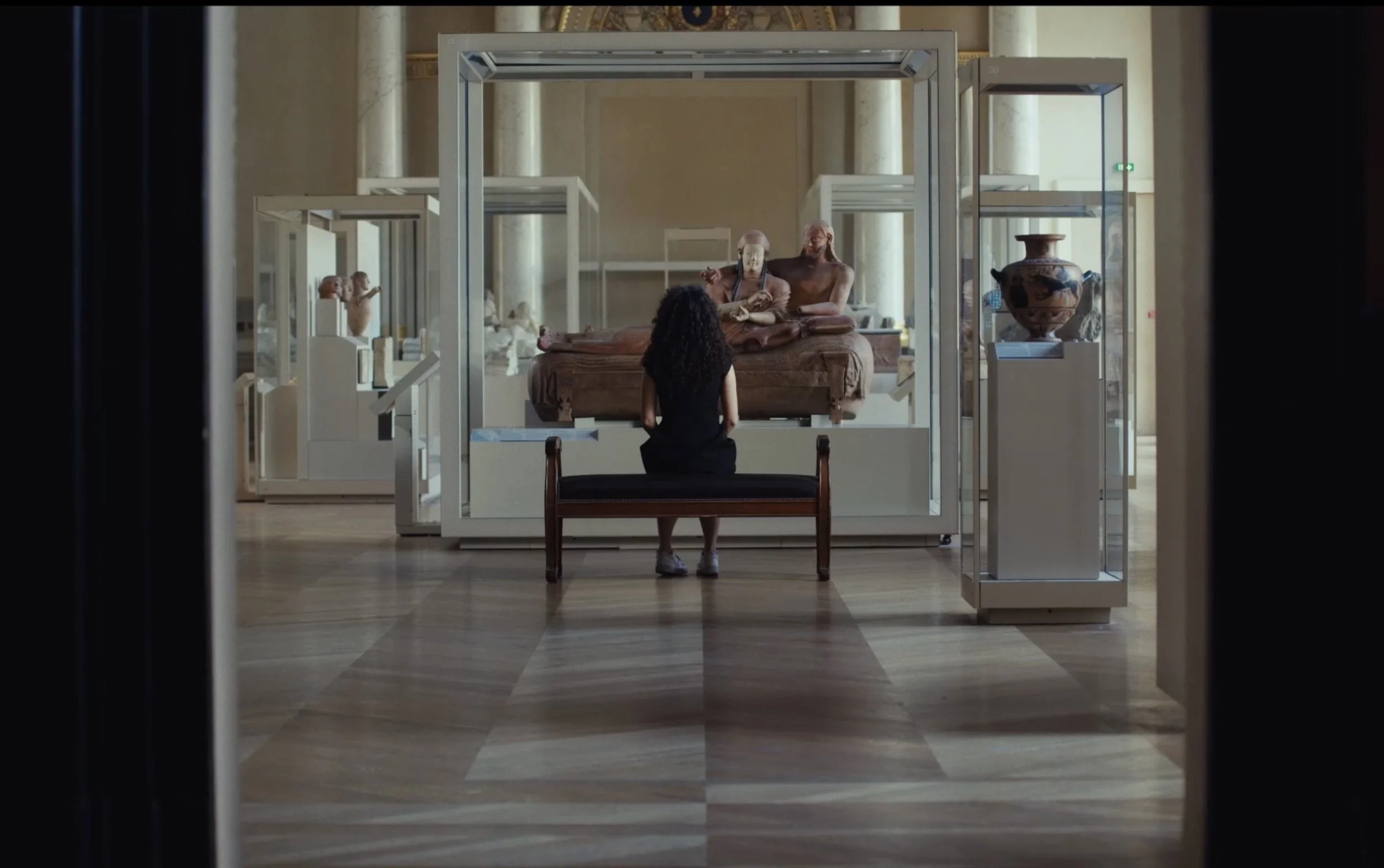 Une femme assise sur un banc devant une vitrine d'exposition dans un musée, regardant une sculpture antique sur un piédestal.