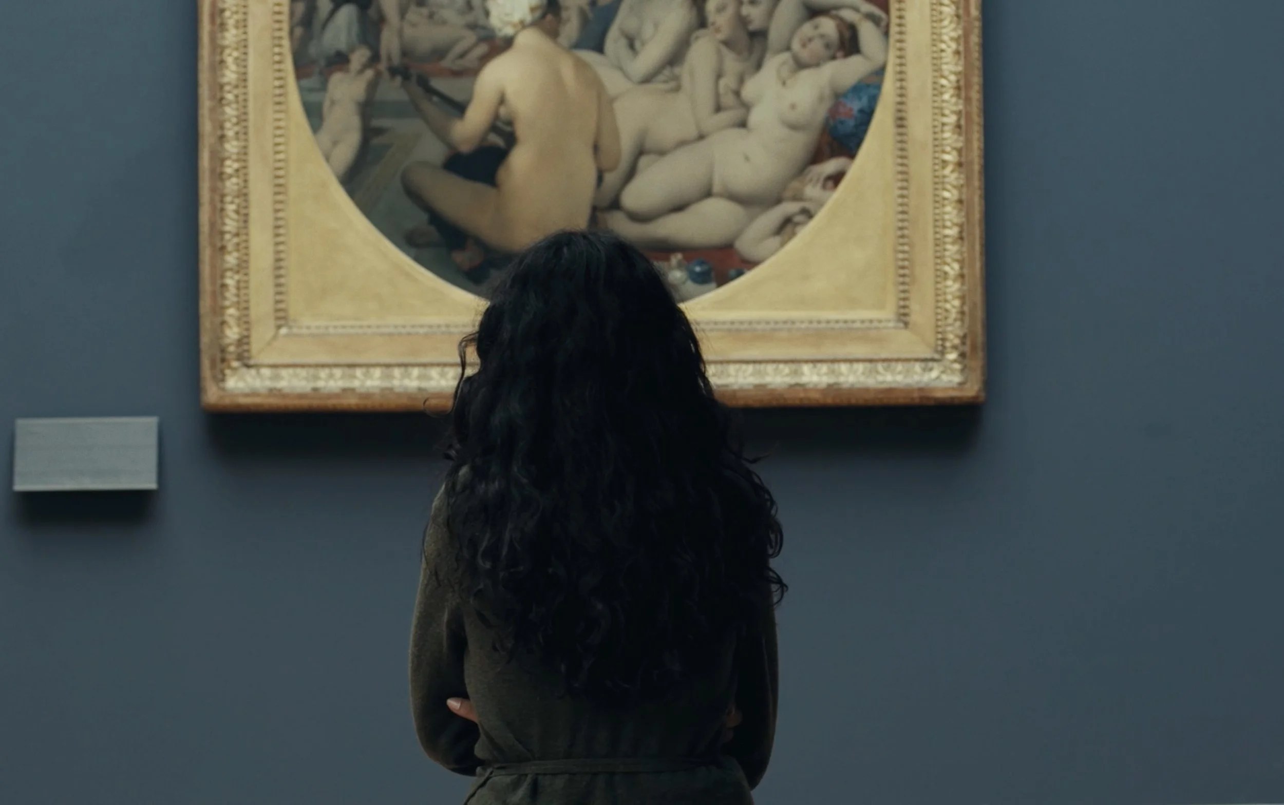 Une femme cheveux longs regarde un tableau représentant des figures nues d'hommes et de femmes dans un musée.