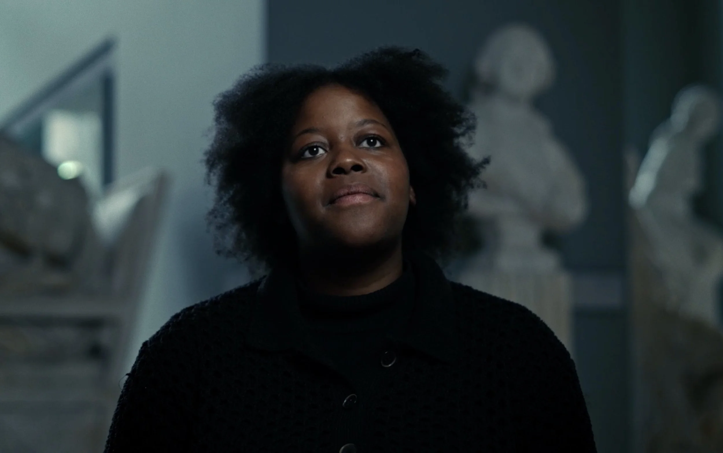 Une femme noire avec des cheveux bouclés regarde vers le haut dans une pièce avec des sculptures en marbre en arrière-plan.