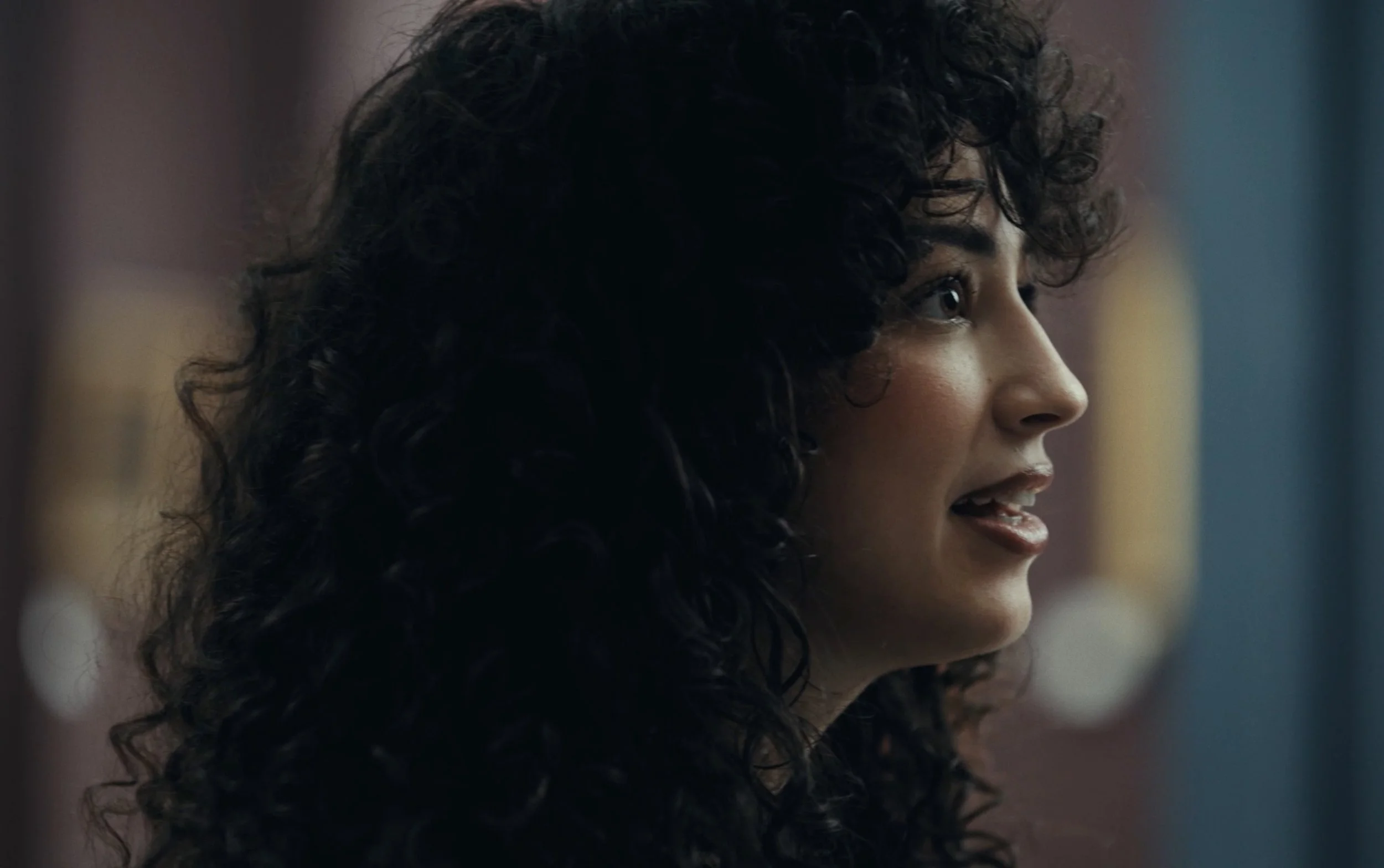 Profil d'une femme avec des cheveux bouclés foncés, souriante, en intérieur avec fond flou.