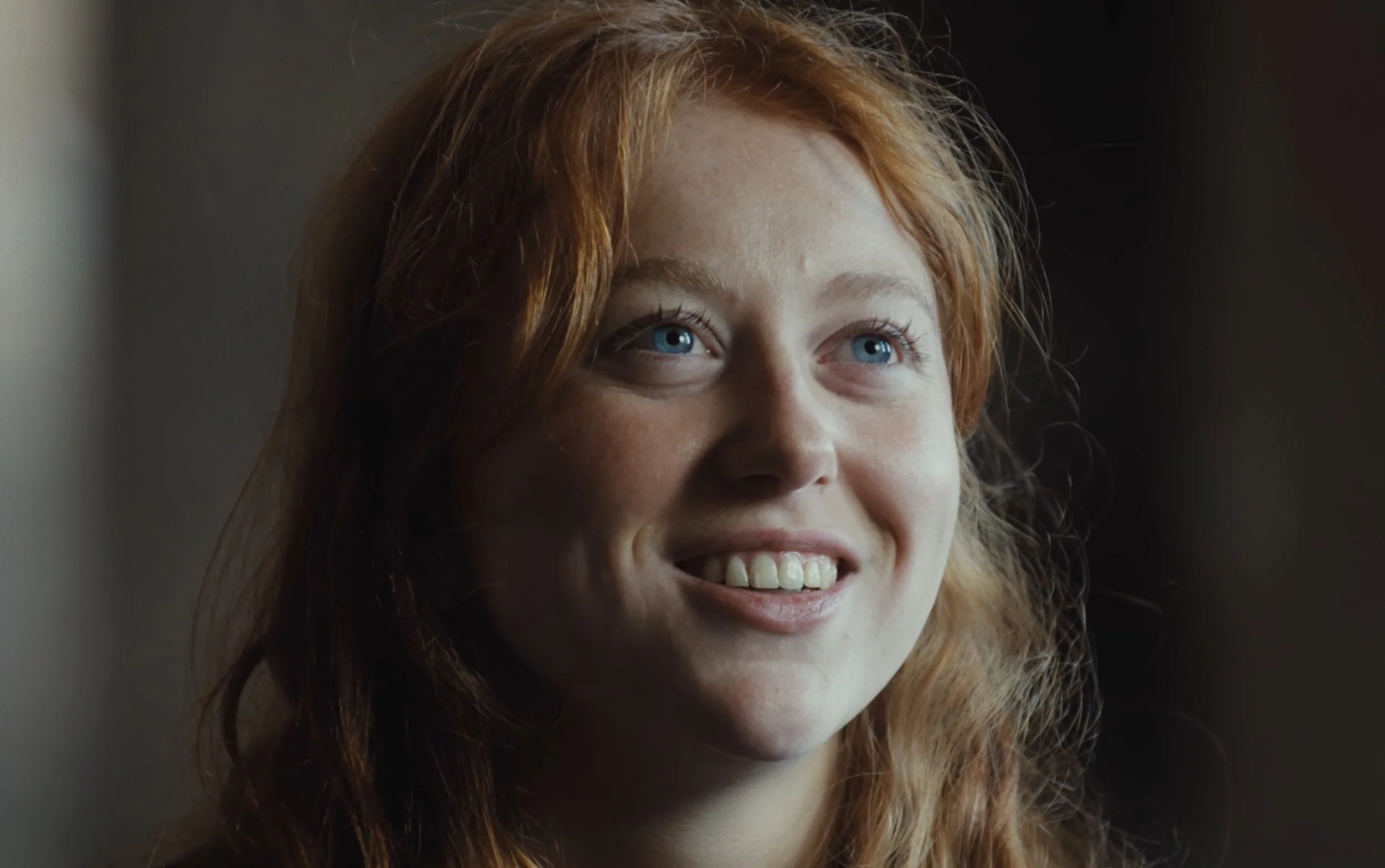 Jeune femme aux cheveux roux et aux yeux bleus souriant, éclairée par une lumière douce.