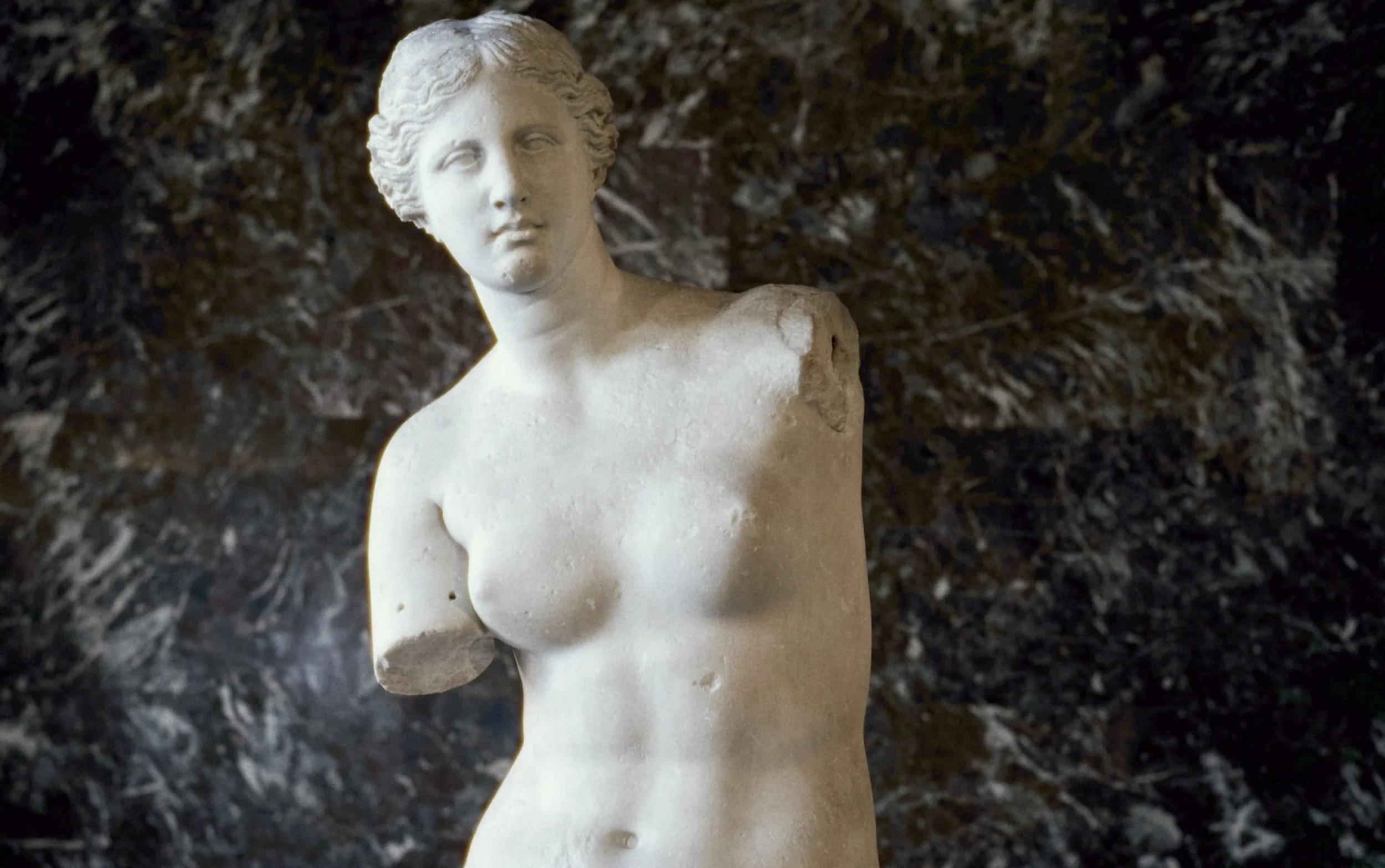 Sculpture en marbre d'une femme nue, cassée au niveau du bras droit, avec un fond sombre et rocheux.