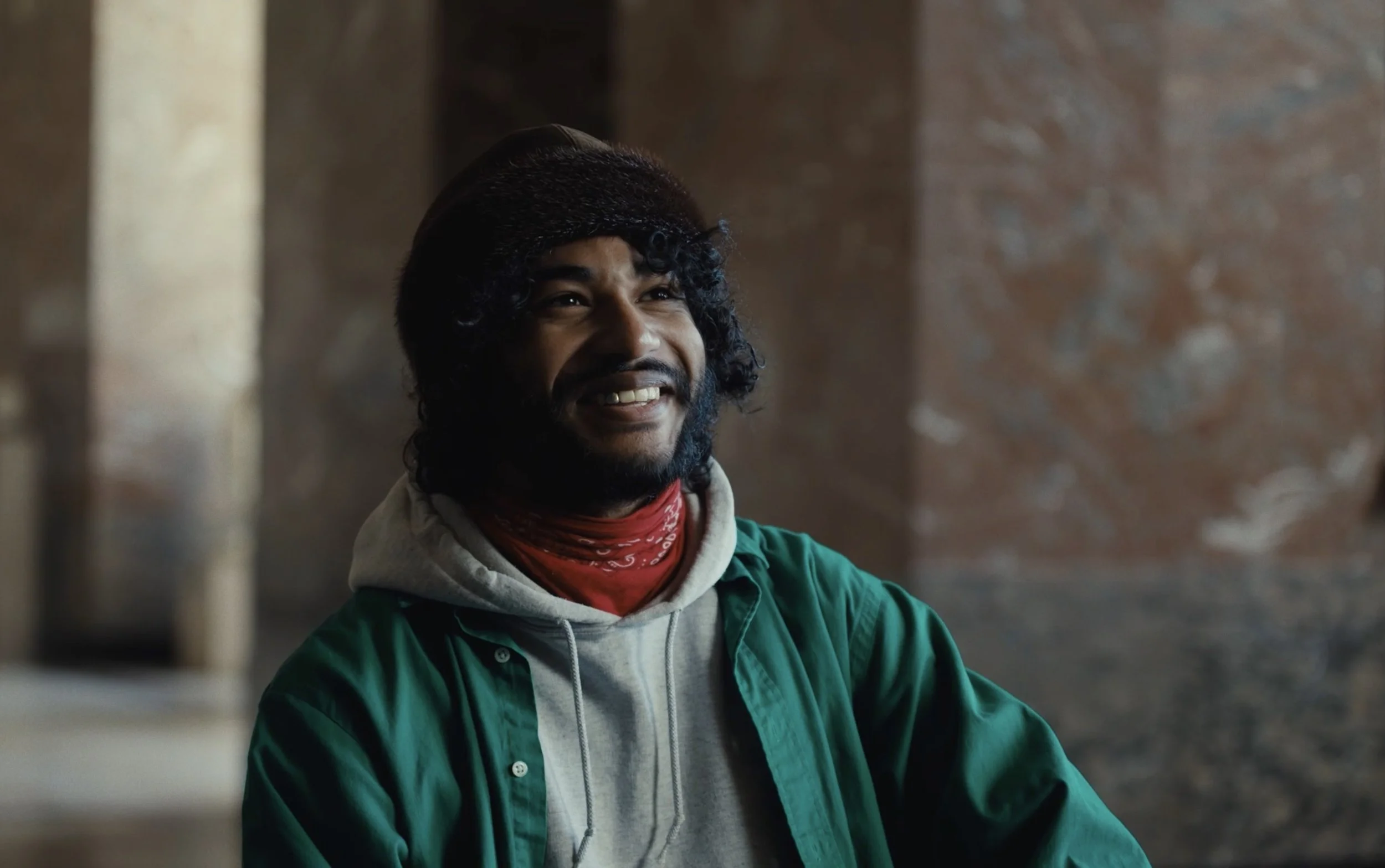 Un homme souriant avec une barbe et des cheveux bouclés, portant un sweat à capuche gris, une chemise verte par-dessus, et un bandana rouge, dans un lieu intérieur avec des murs en pierre.