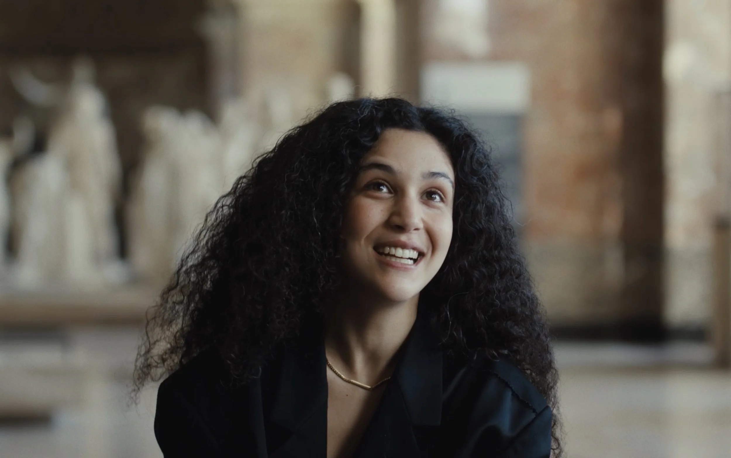 Jeune femme à cheveux bouclés, souriante, en intérieur, portant un vêtement noir, arrière-plan de style industriel avec des sculptures en céramique.