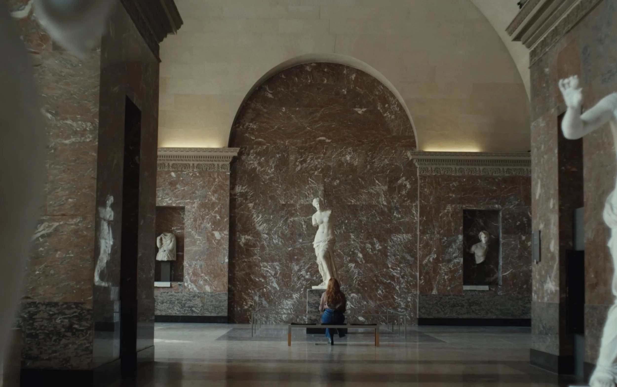 Une femme assise devant une statue de marbre dans un musée, entourée de murs en marbre marron avec des bustes en marbre.