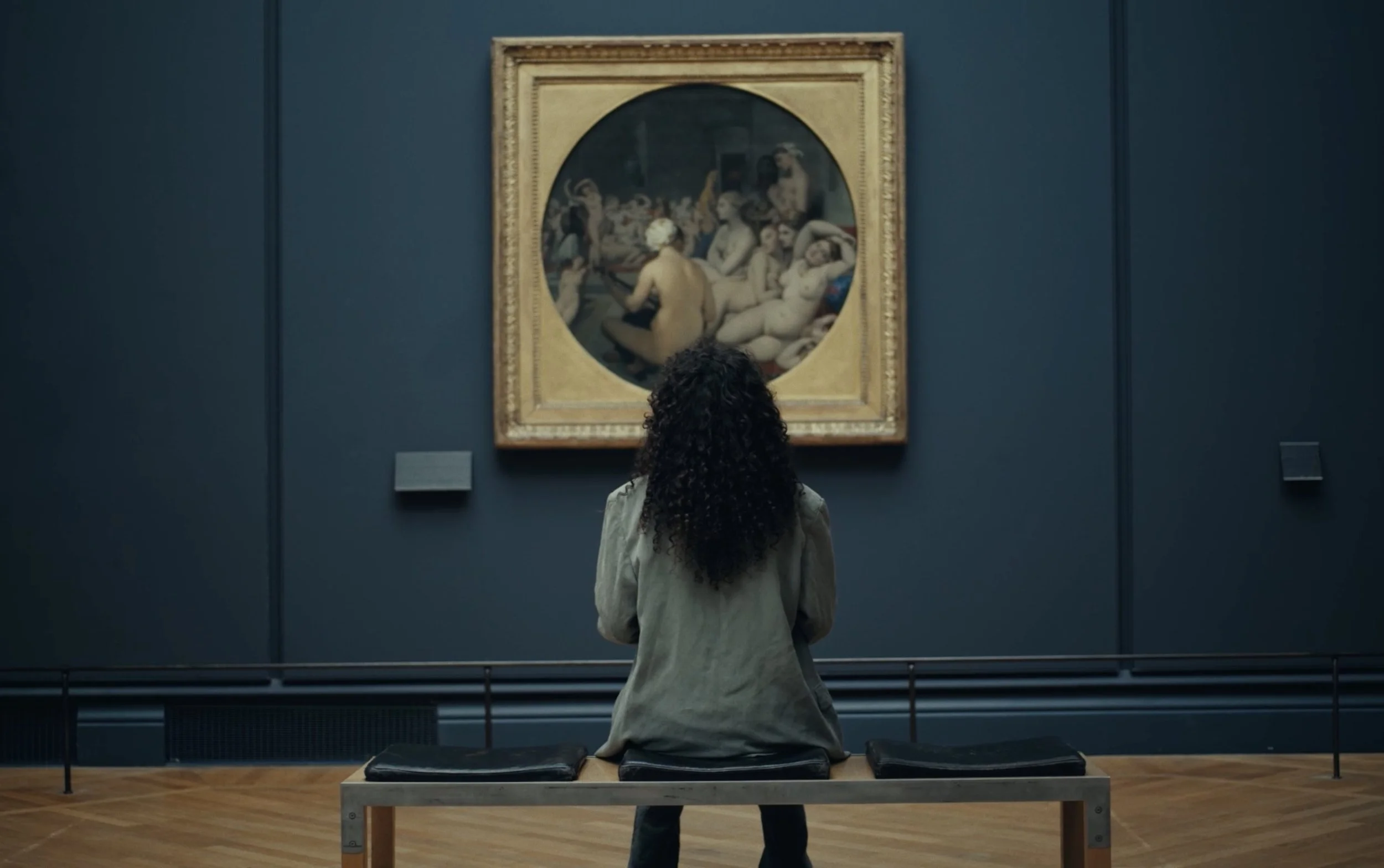 Une femme avec des cheveux bouclés assise sur un banc, regardant un tableau dans un musée, représentant des figures nues.