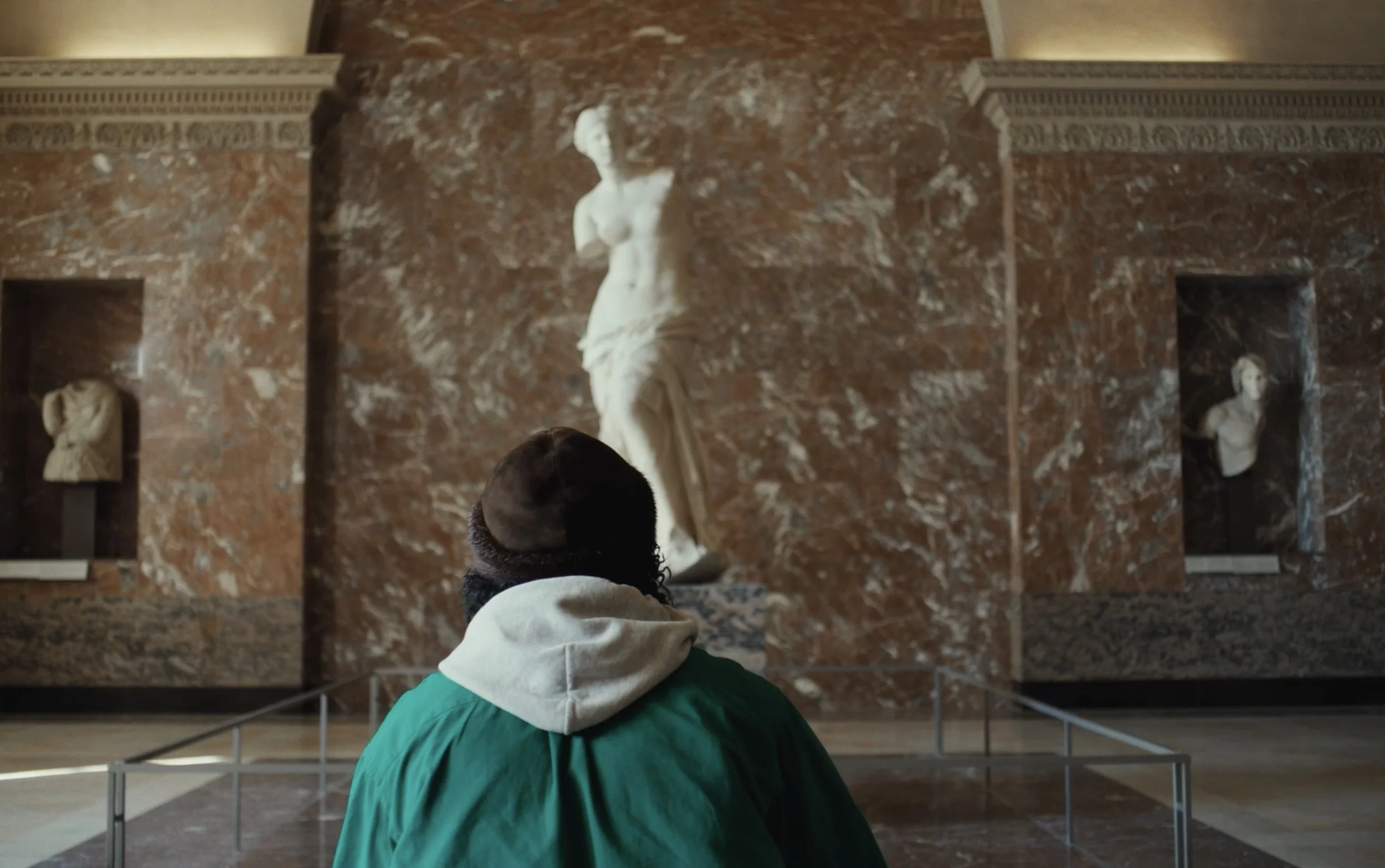 Une personne vue de derrière regarde une sculpture de la Vénus de Milo dans un musée, avec un mur en marbre derrière.