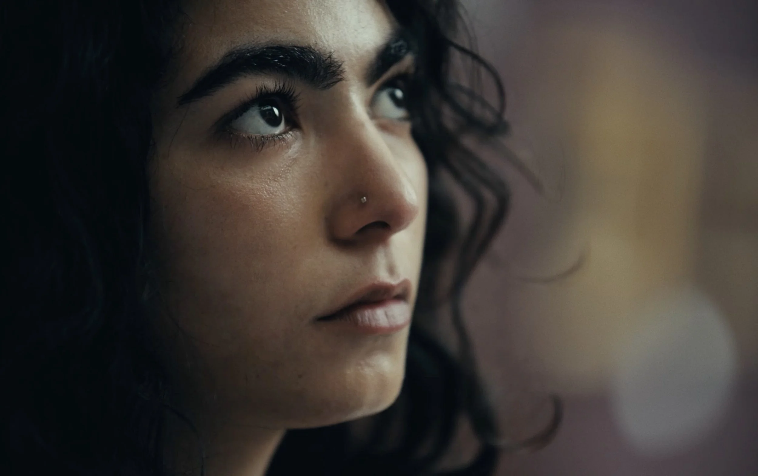 Close-up d'une femme avec des cheveux bouclés noirs, regardant vers la gauche, avec un maquillage naturel et un piercing au nez.
