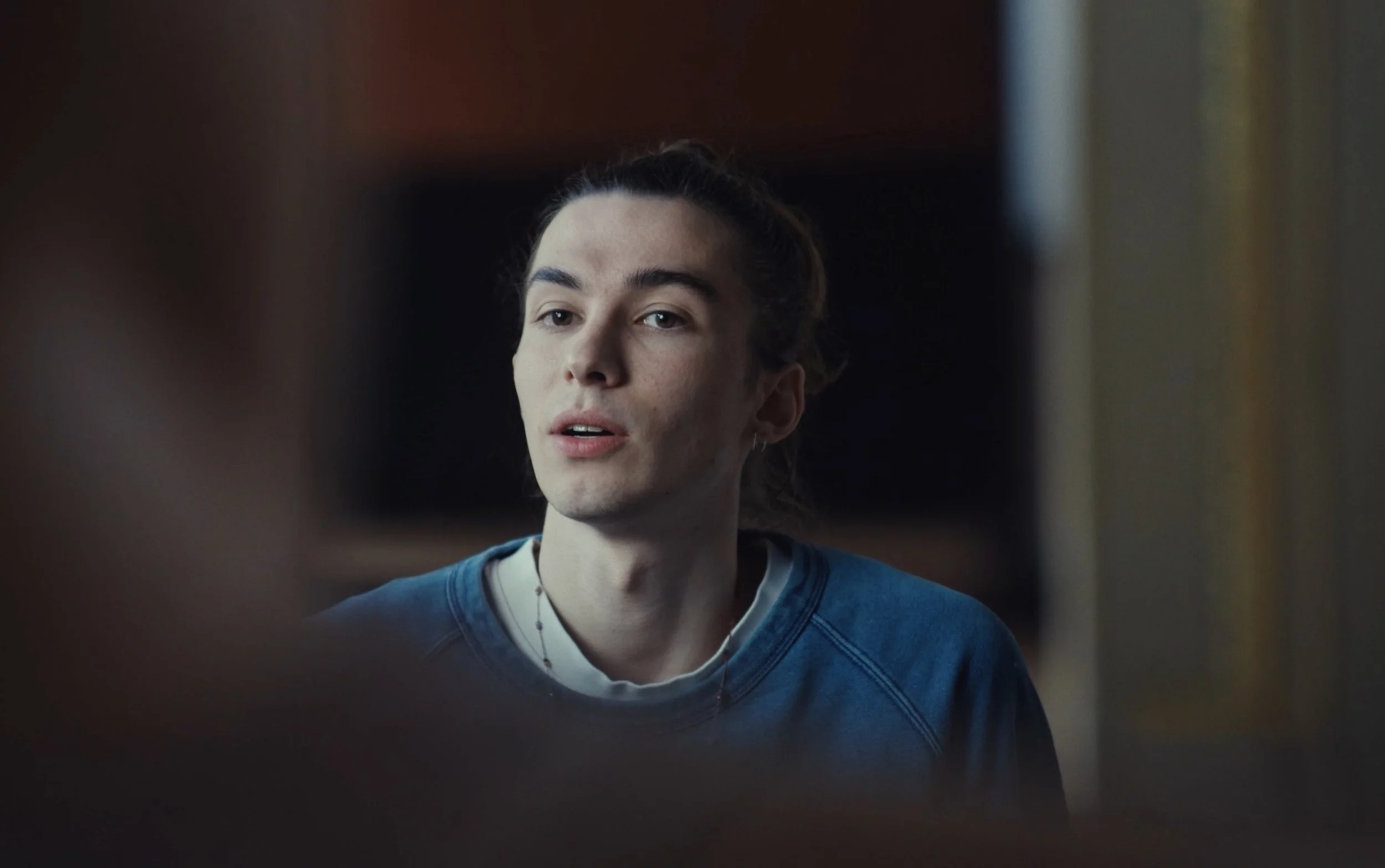 Un jeune homme avec des cheveux attachés, portant un sweat bleu, regarde légèrement vers la gauche dans une pièce. Le fond est sombre et flou.