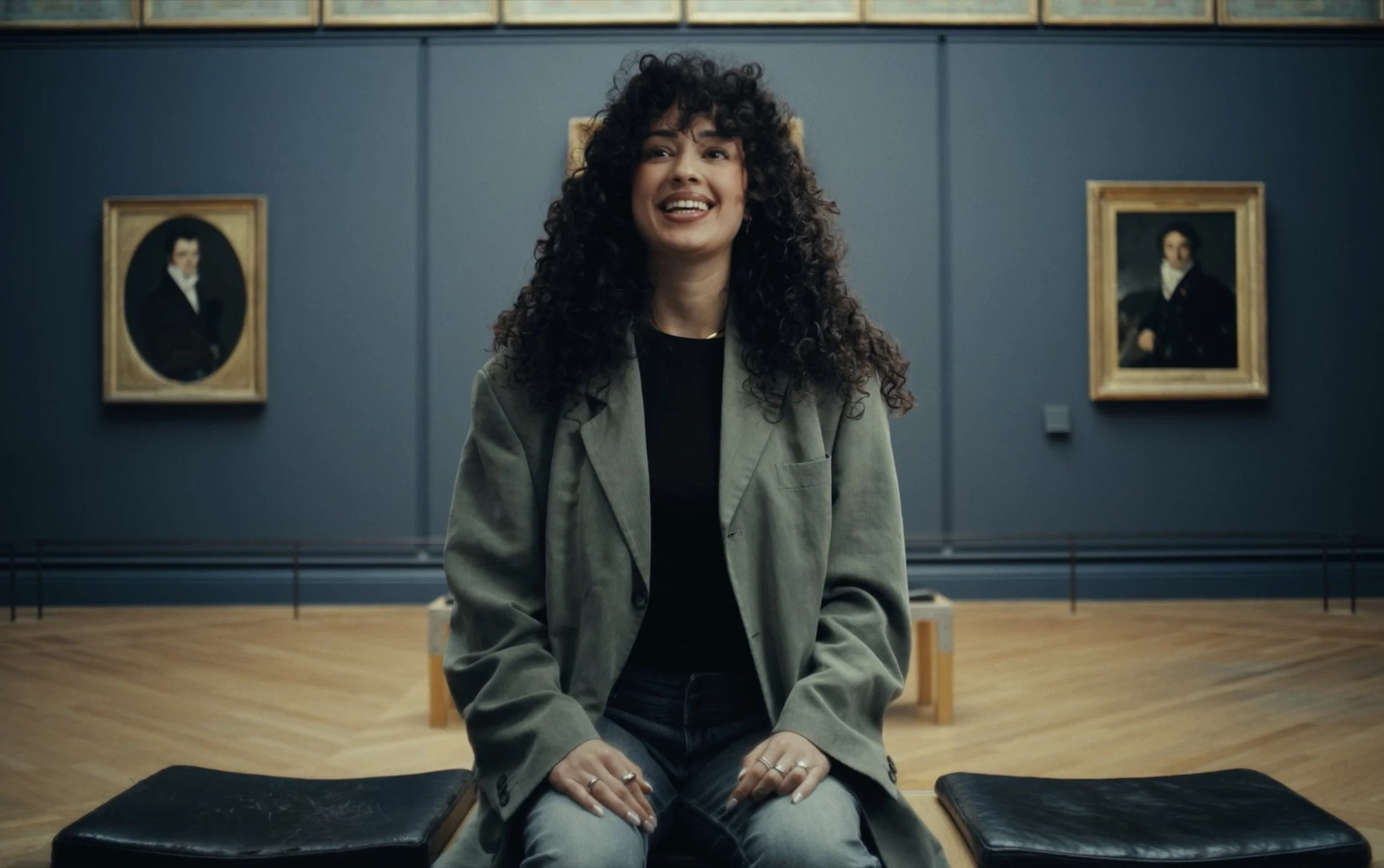 Une femme assise dans un musée, souriante, avec deux portraits classiques en arrière-plan.