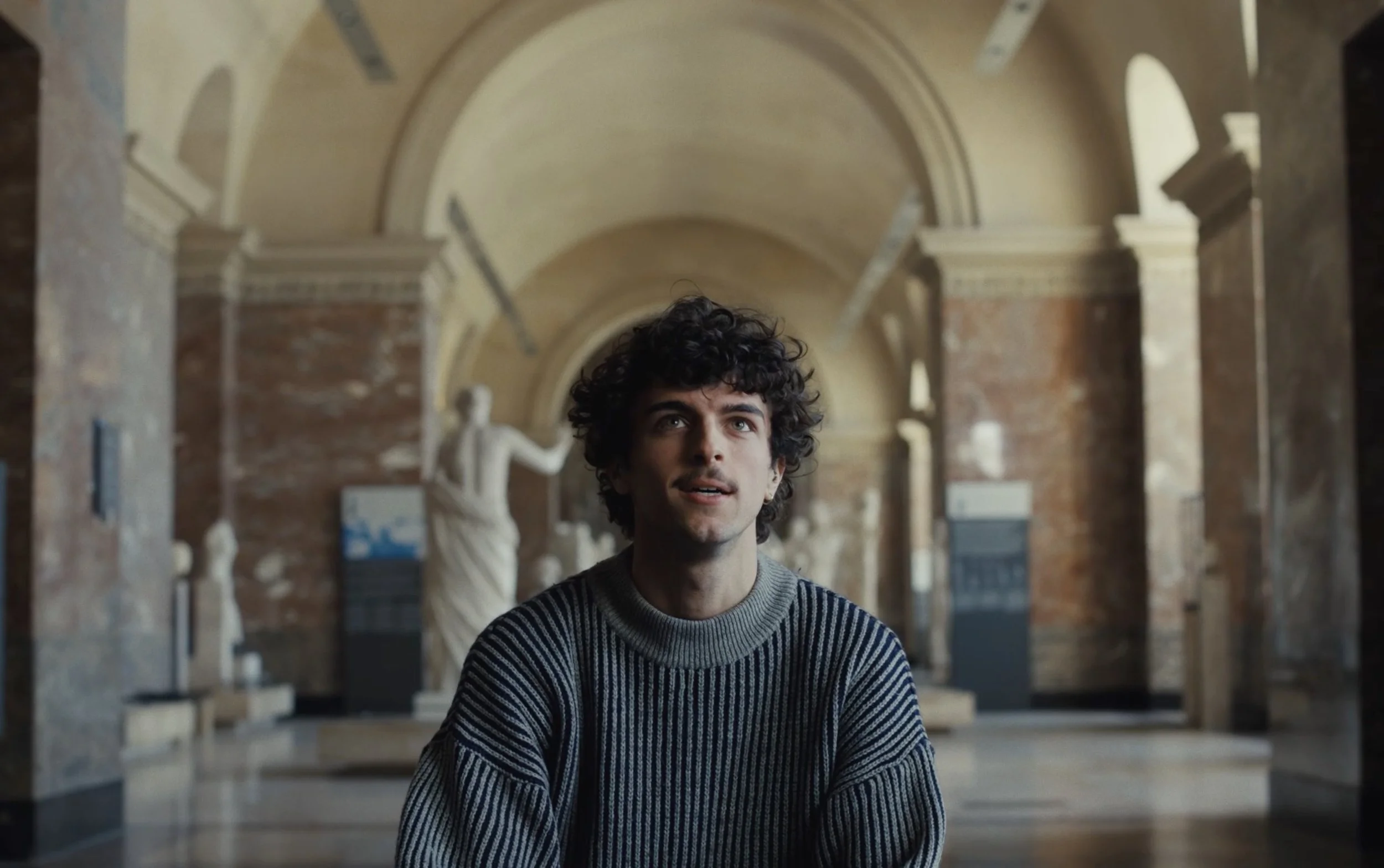 Jeune homme aux cheveux bouclés, portant un pull rayé gris, assis dans un musée avec des sculptures classiques en arrière-plan.