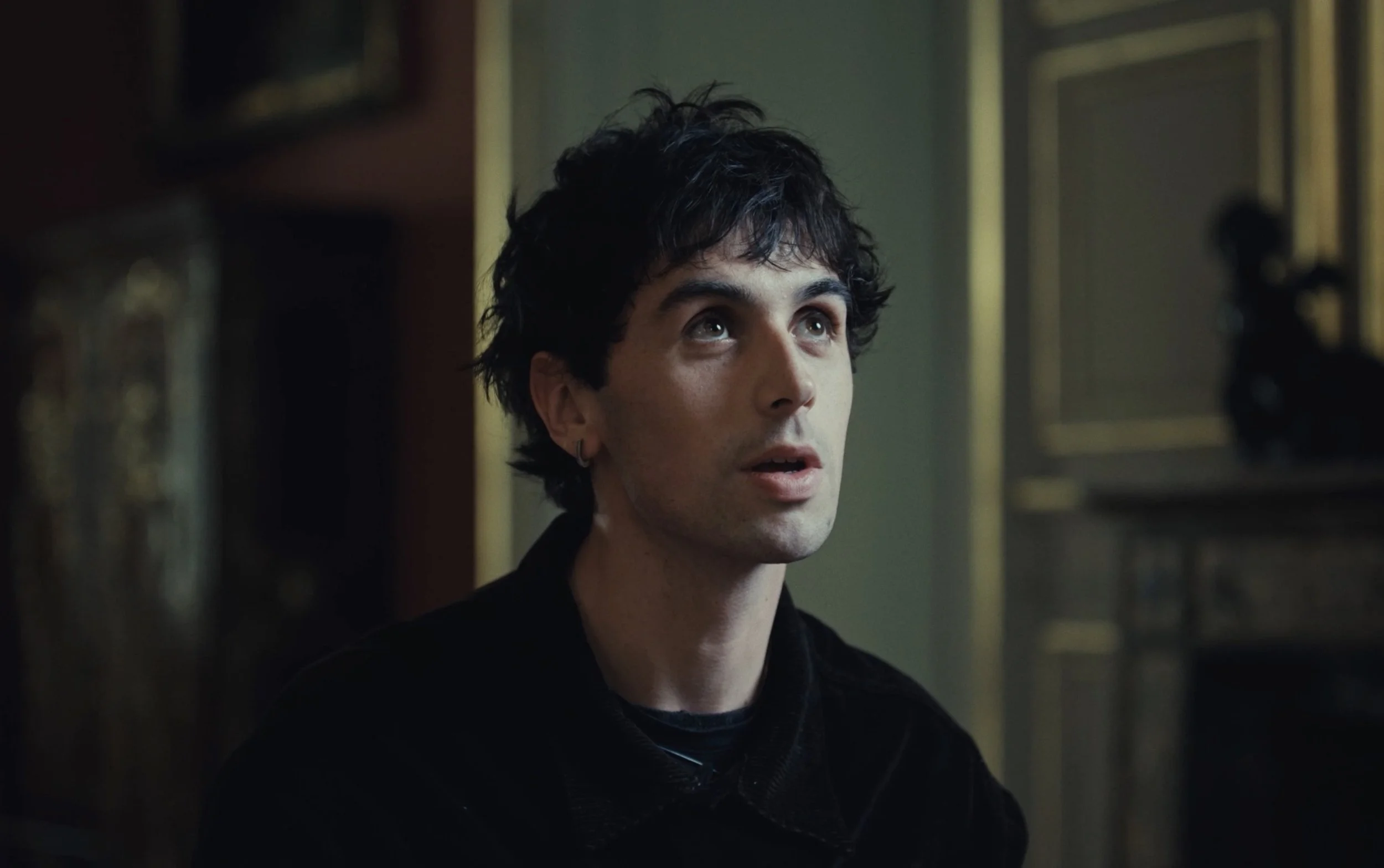 Un homme avec des cheveux noirs bouclés et un petit anneau à l'oreille regarde vers le haut dans une pièce décorée d'œuvres d'art et de moulures dorées.