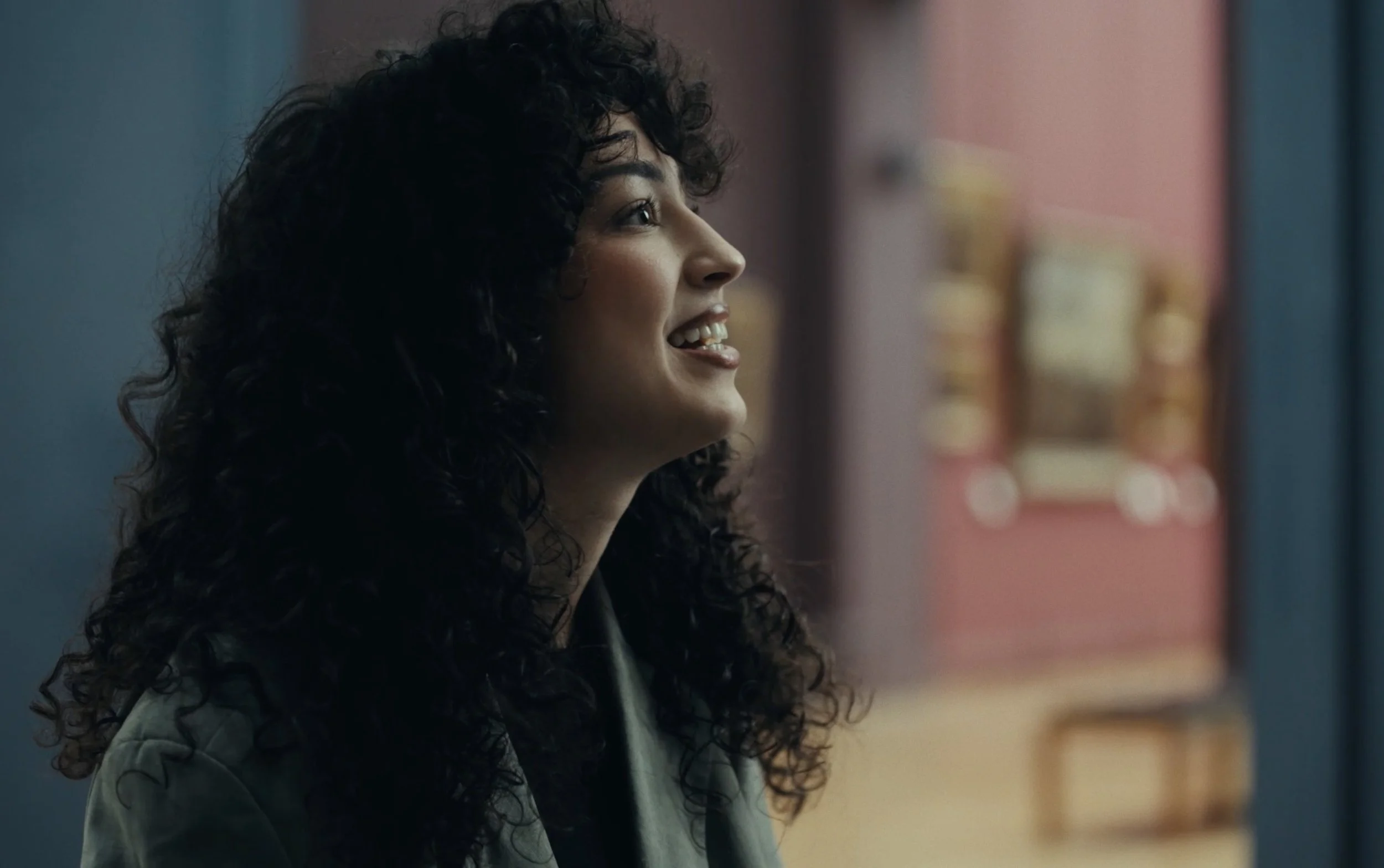 Femme avec cheveux bouclés foncés, souriante, regardant vers la droite, dans un intérieur avec murs colorés.