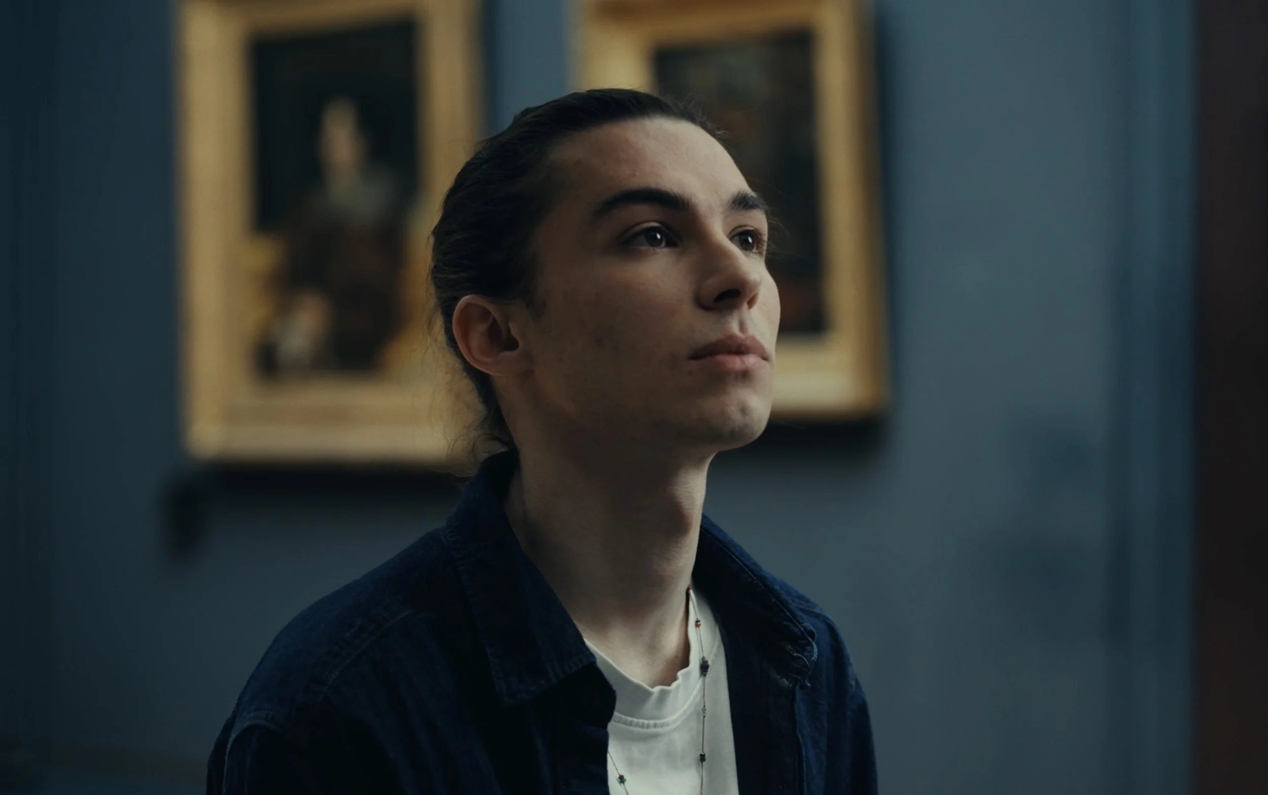 Un jeune homme aux cheveux longs, portant une chaîne et un t-shirt blanc, regarde attentivement vers la droite dans une pièce avec des tableaux en arrière-plan.