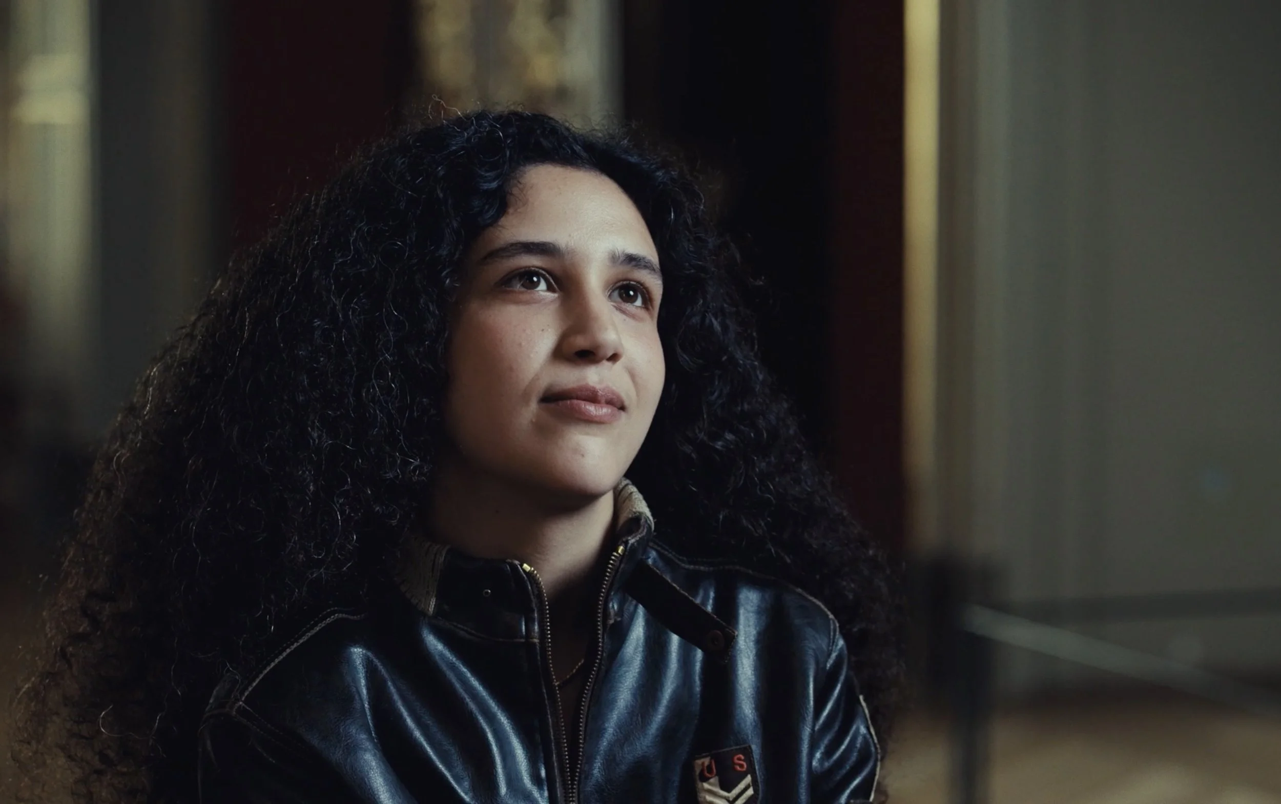 Jeune femme rousse avec de longs cheveux bouclés, portant une veste en cuir noire, assise, regardant légèrement vers le haut dans un intérieur élégant.
