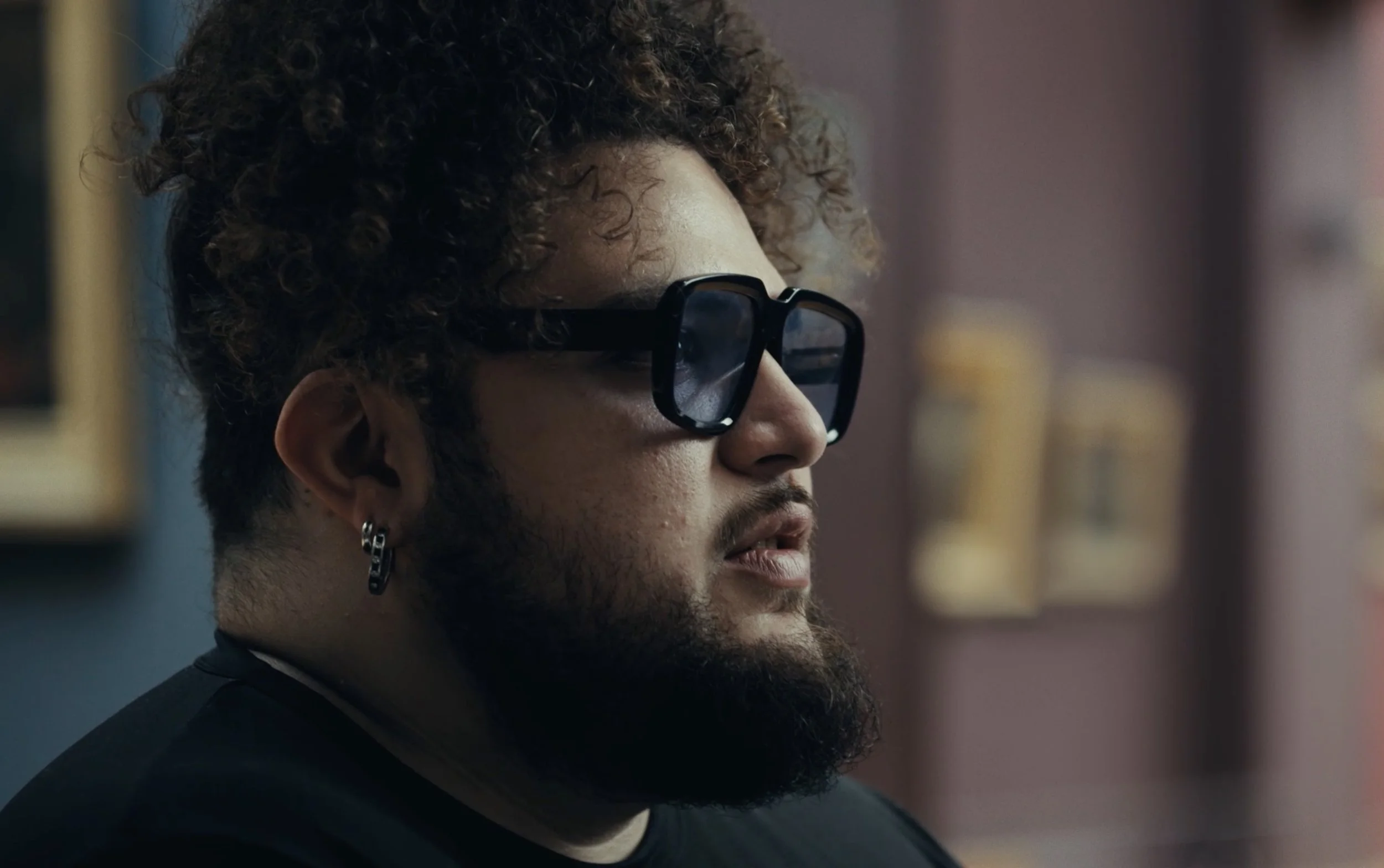 Un homme avec des lunettes de soleil, une barbe et des boucles dans les cheveux, portant un earring en cercle.