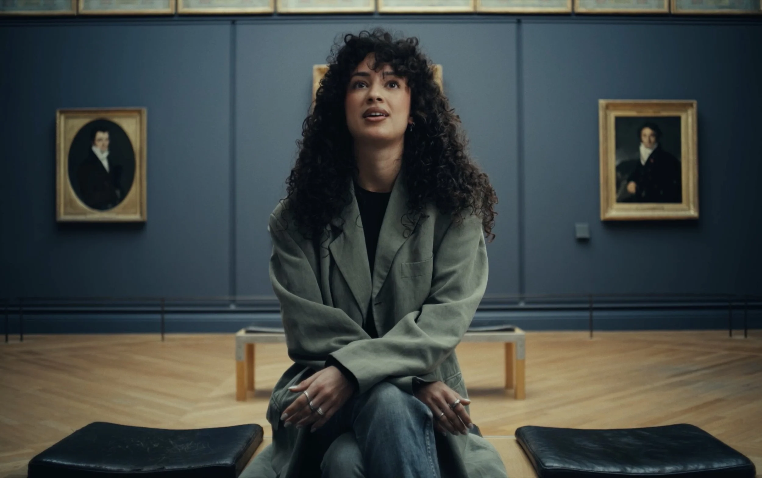 Une femme aux cheveux bouclés assise dans un musée, avec des portraits encadrés en arrière-plan.