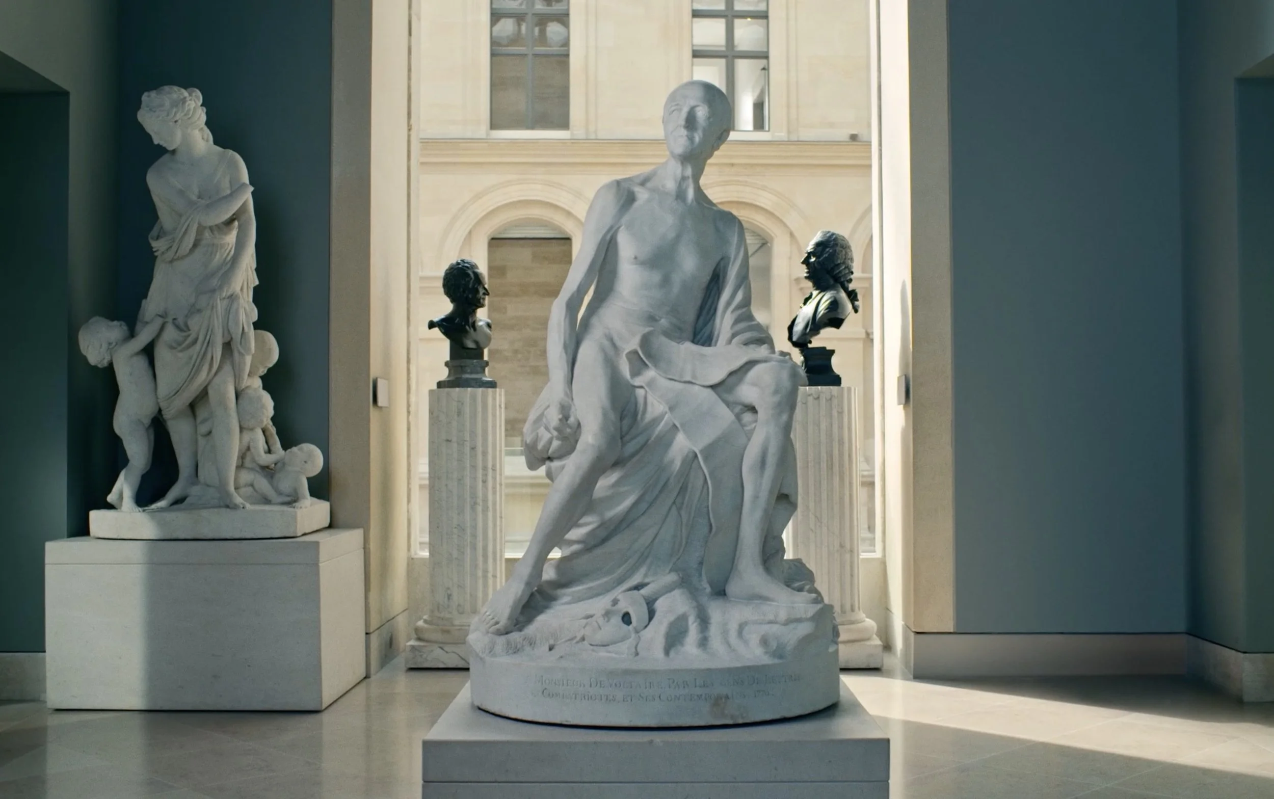 Sculpture en marbre représentant une figure humaine assise, située dans un musée, avec d'autres bustes et sculptures en arrière-plan.