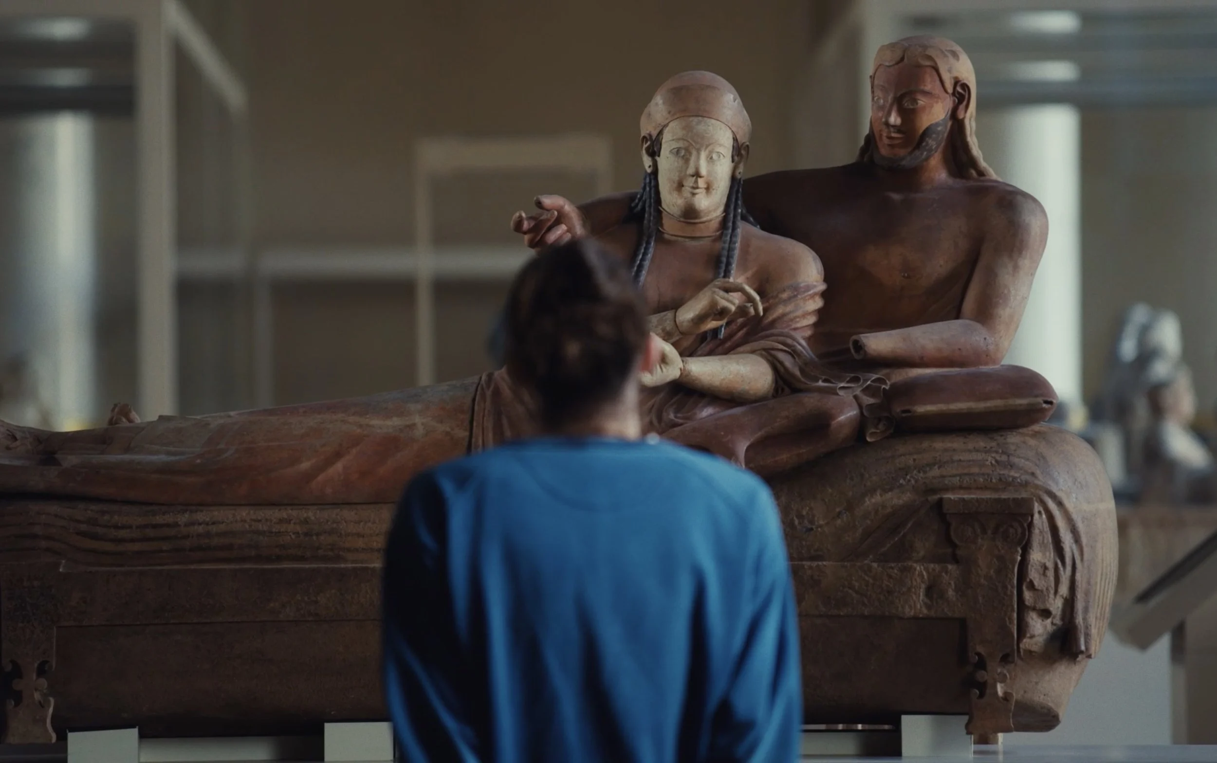 Une personne regarde une sculpture ancienne de deux figures assises, exposée dans un musée.