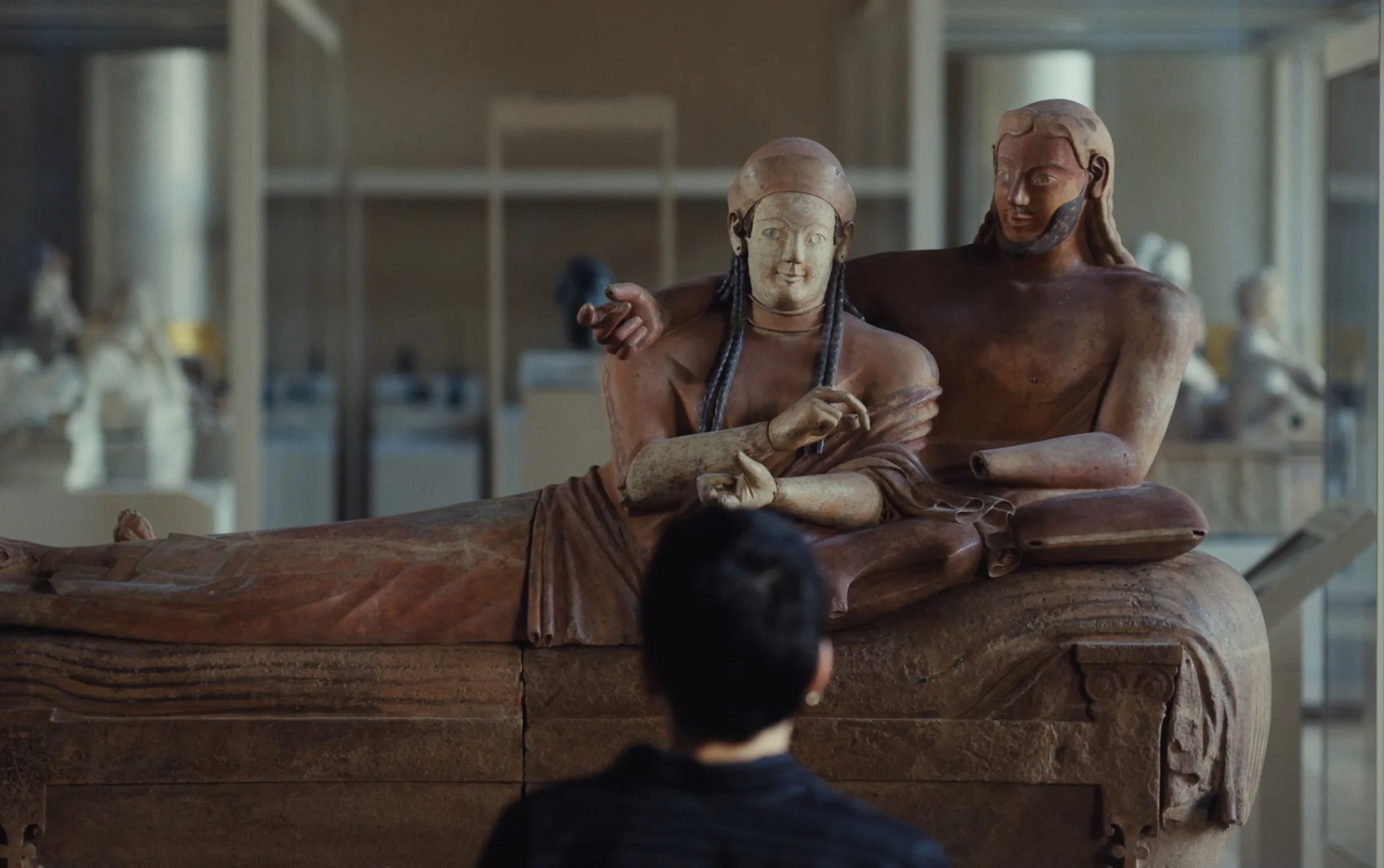 Une personne regarde un sarcophage antique orné de sculptures représentant deux figures mythologiques ou religieuses. La scène est dans un musée.