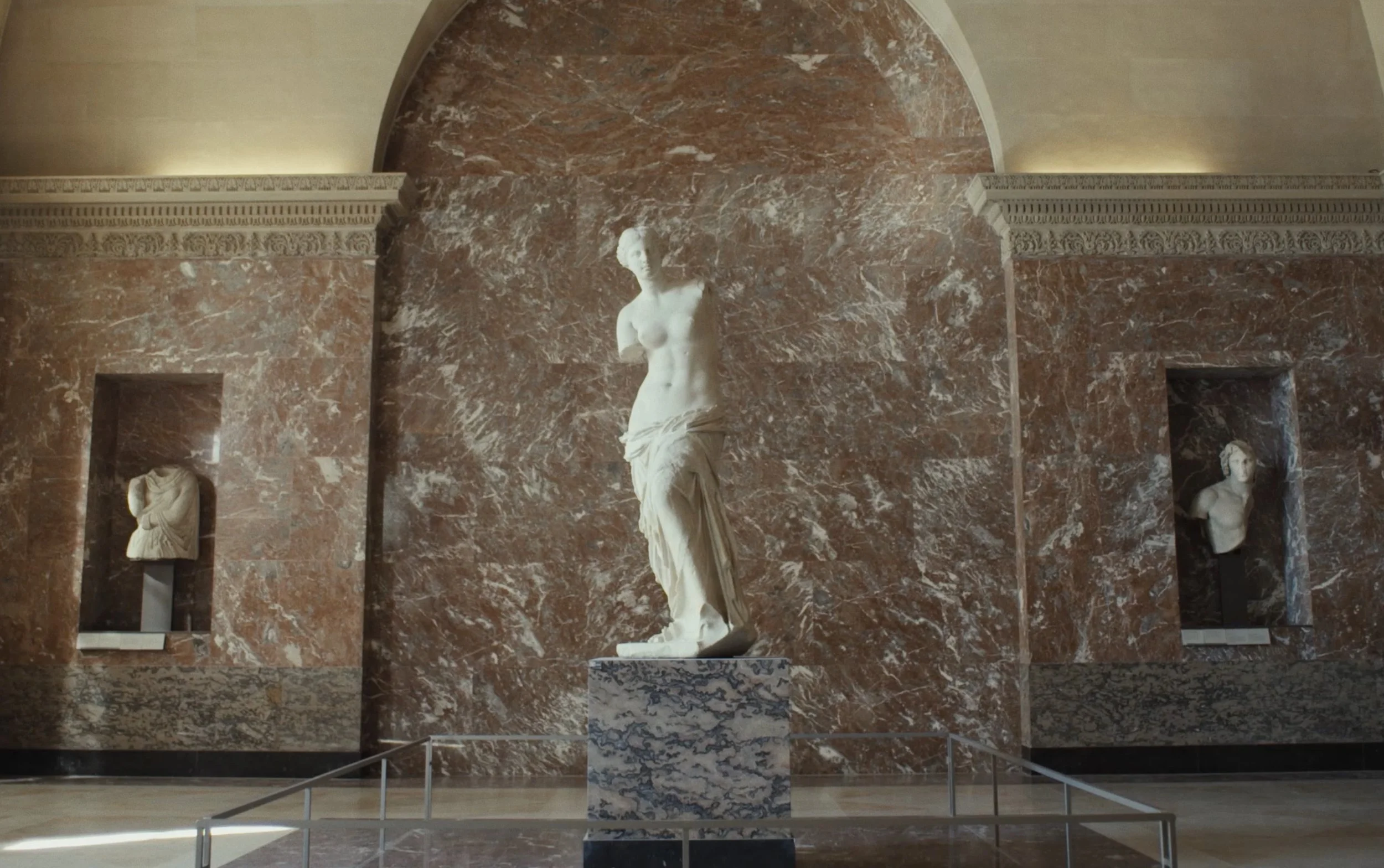 Statue en marbre d'une femme nue, positionnée sur un piédestal en marbre, dans une salle avec murs en marbre rouge et décorations classiques.