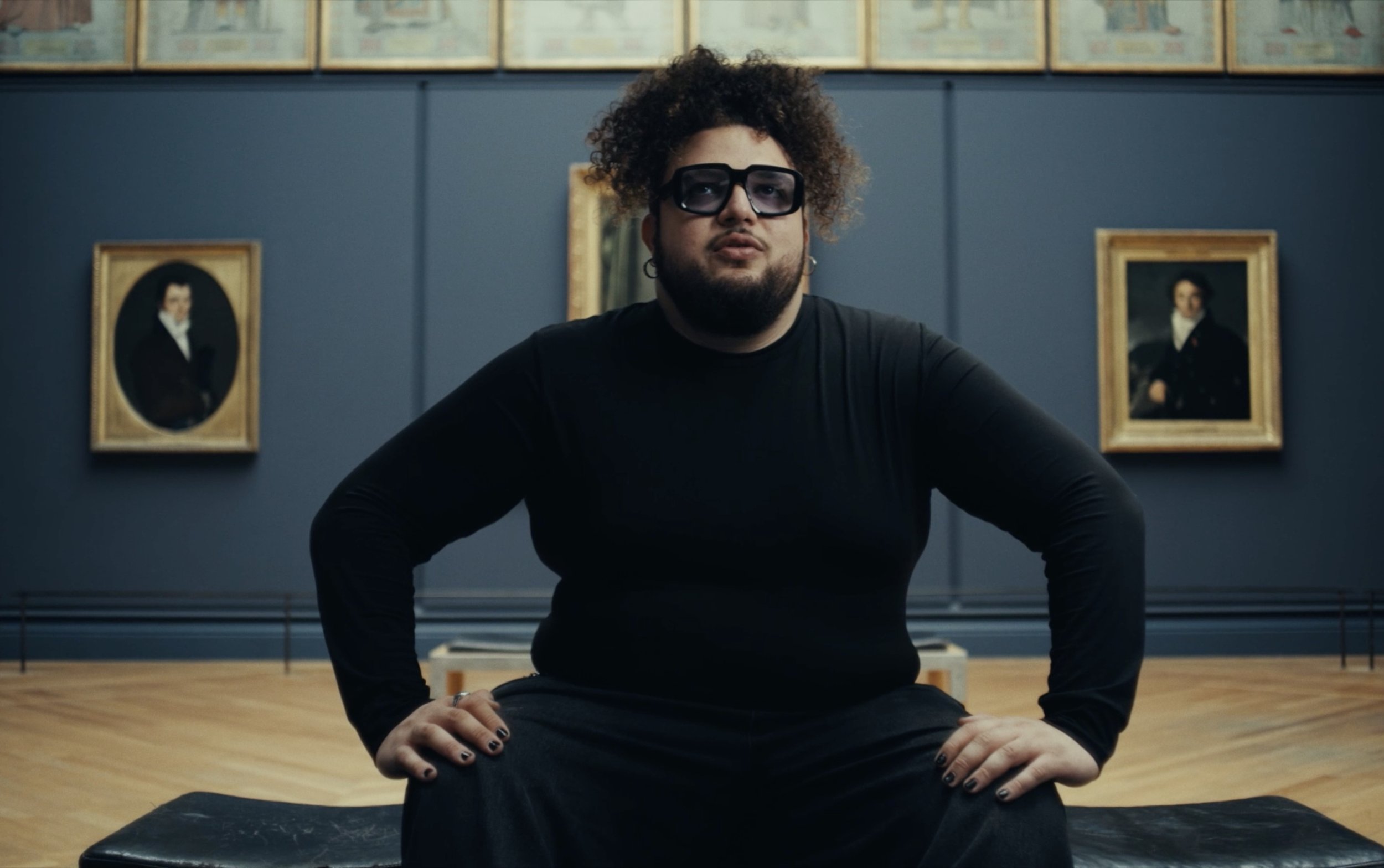 Une personne assise au centre d'un musée, portant des lunettes de soleil et une tenue noire, avec des portraits en peinture en arrière-plan.