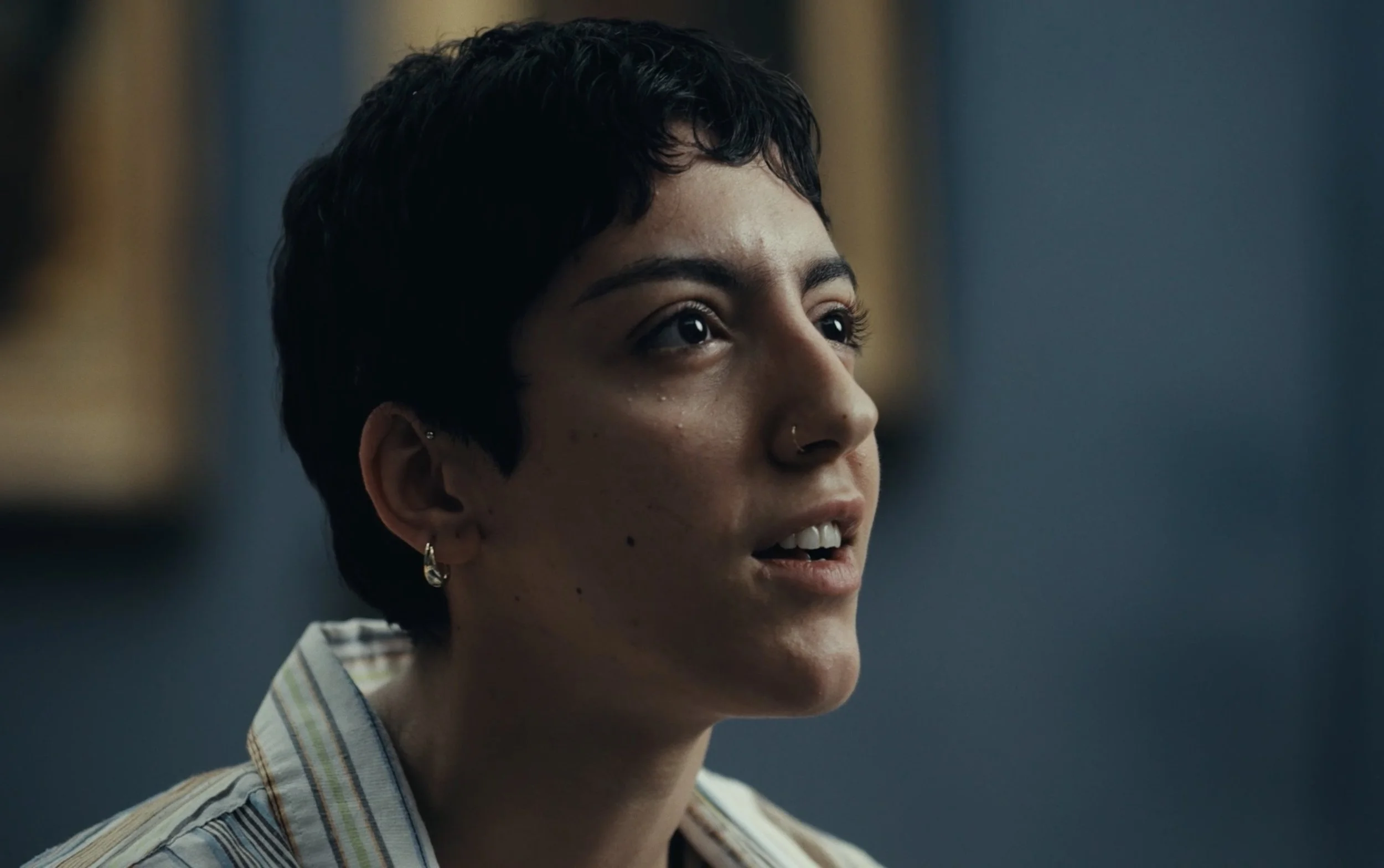 Une jeune femme avec des cheveux courts, portant un piercing au nez et un hoop à l'oreille, regarde légèrement vers la droite, dans un environnement intérieur.