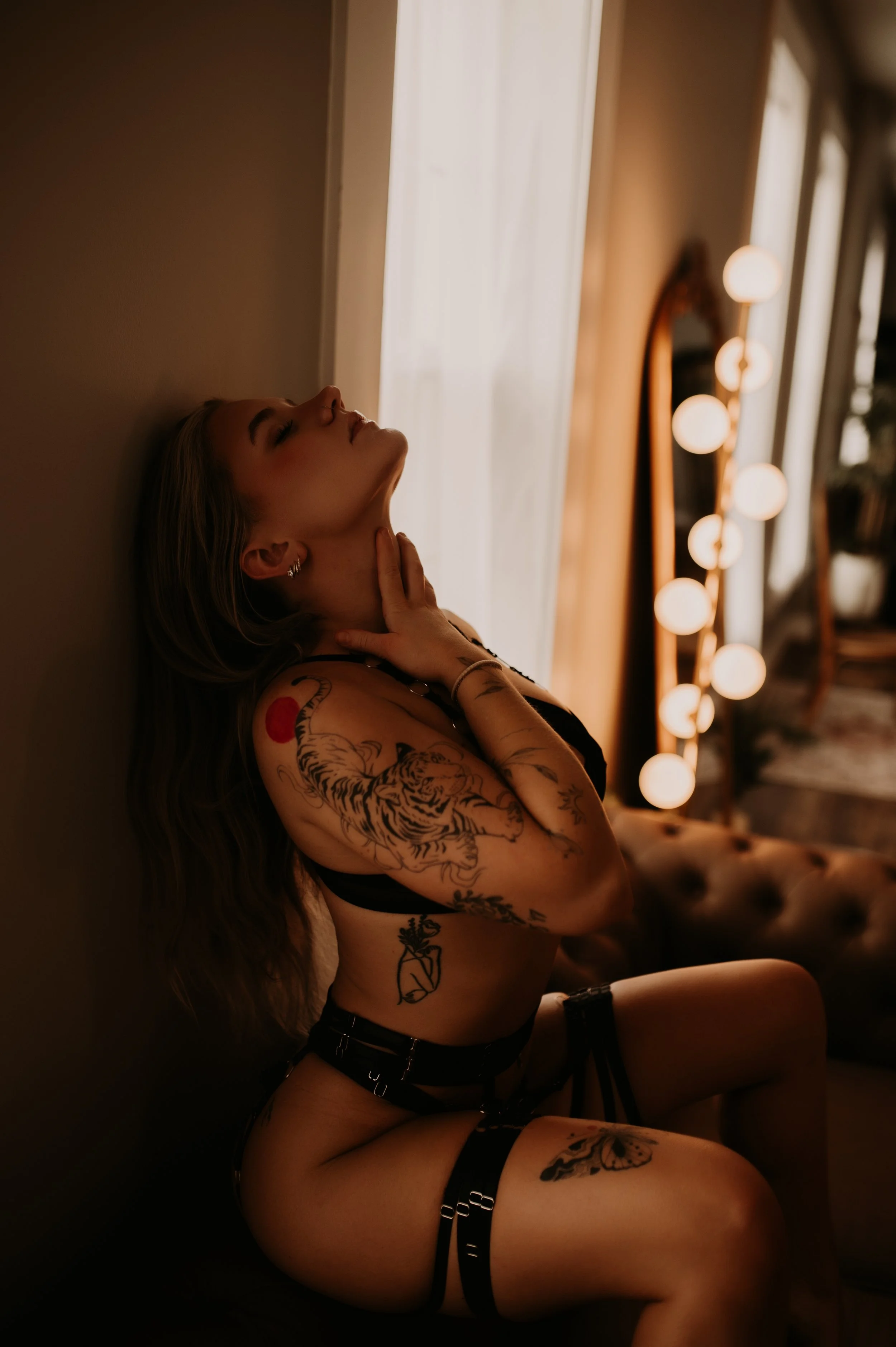 A woman with tattoos on her arms and legs, sitting on a couch with her head tilted back and hand on her neck, in a dimly lit room with a mirror and round lights in the background.