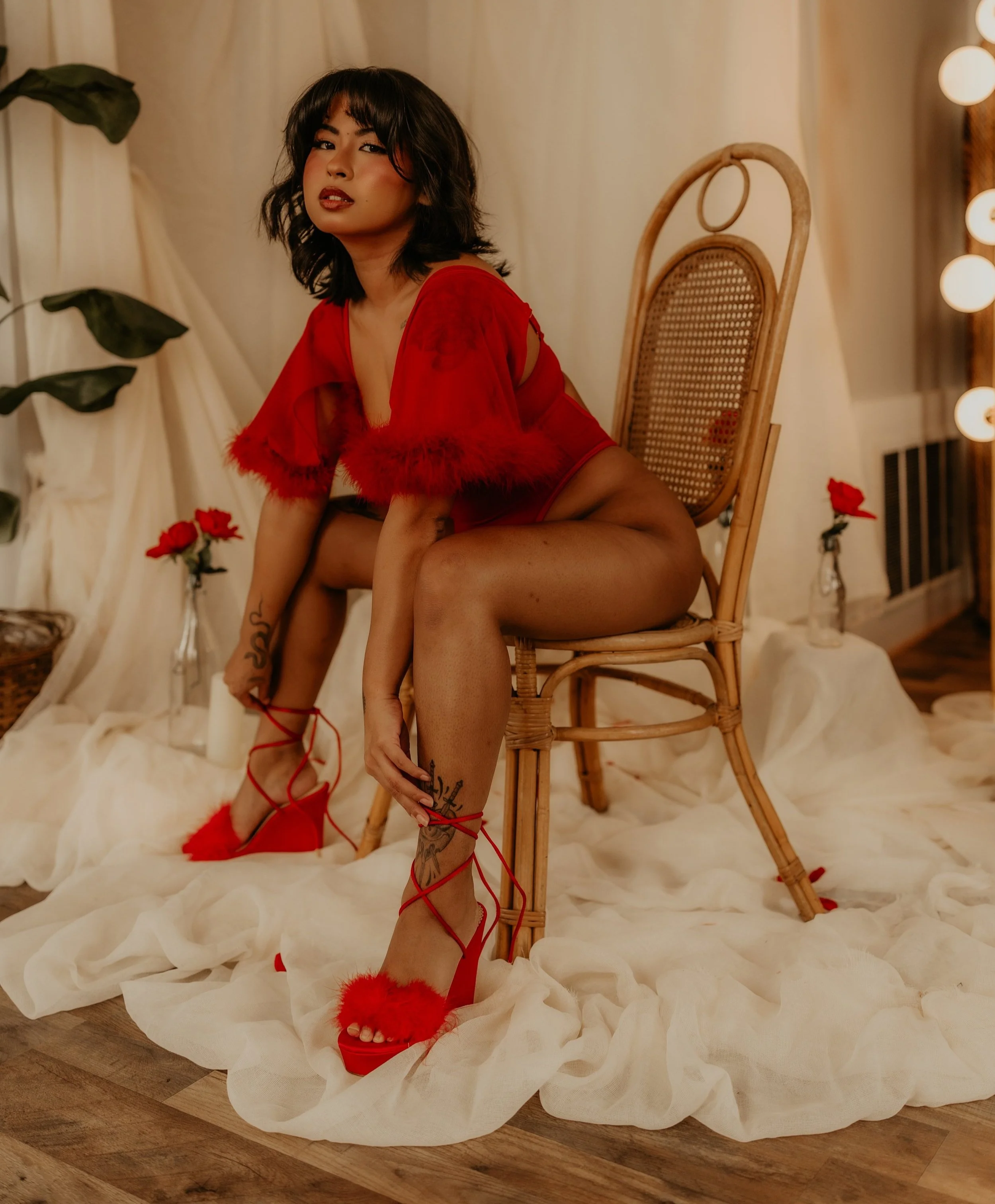 A woman in red lingerie and fluffy red high heels posing on a wooden floor surrounded by white fabric, with roses and warm lighting in a cozy setting.