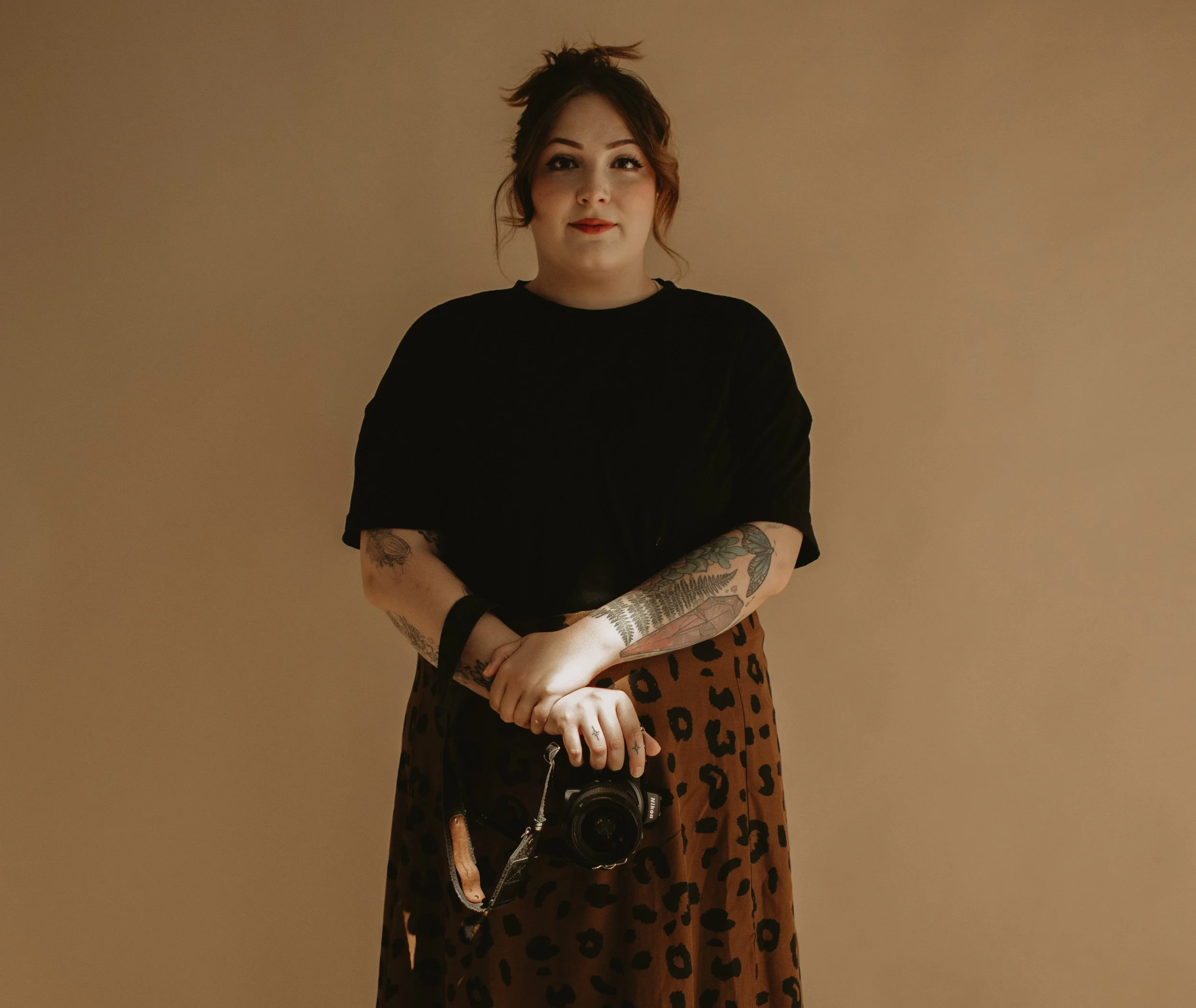 A woman with tattoos holding a camera, wearing a black top and a leopard print skirt, standing against a plain beige background.