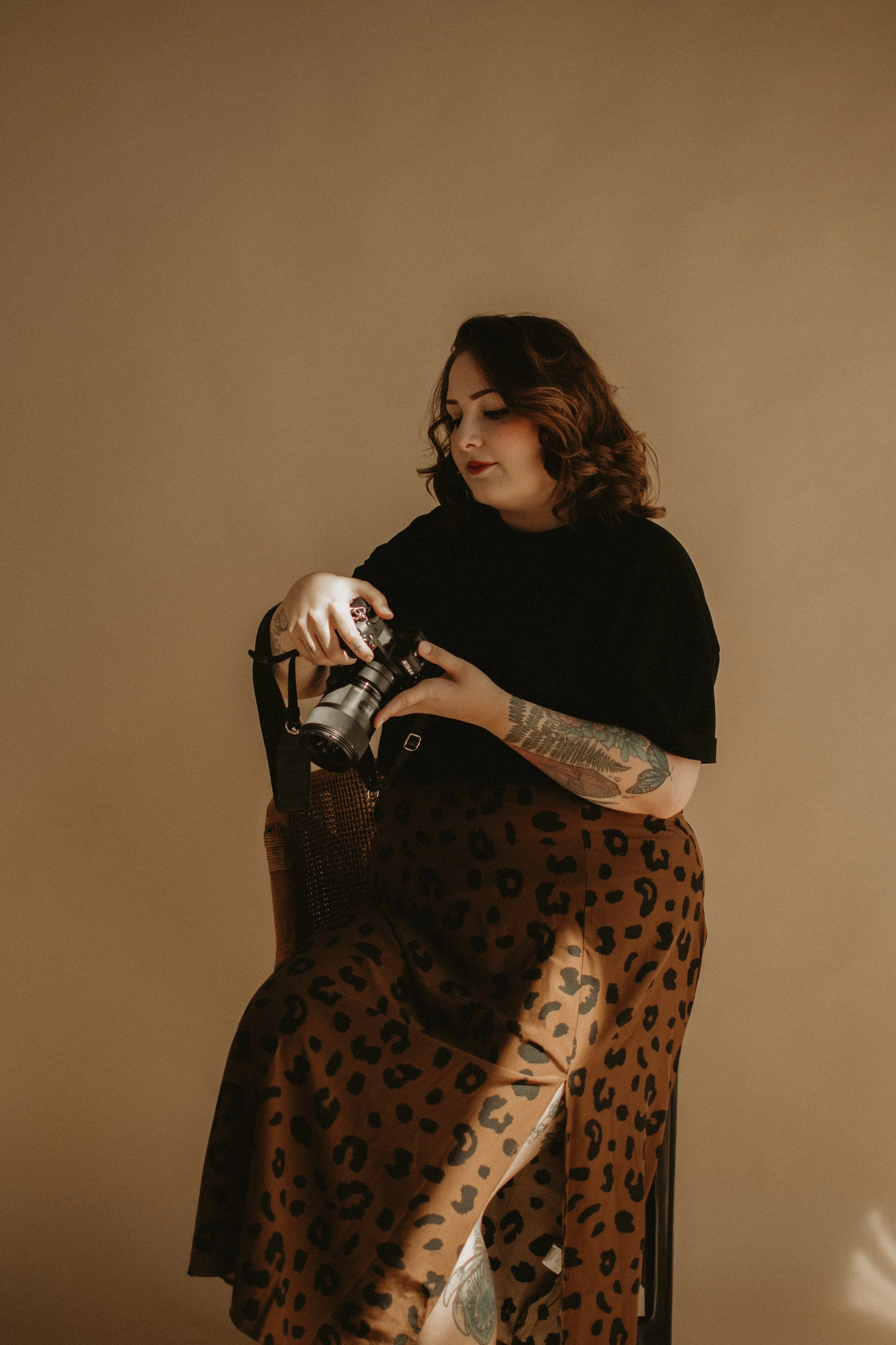 A woman with curly hair wearing a black top and leopard print skirt, sitting on a chair and holding a camera, against a beige background.