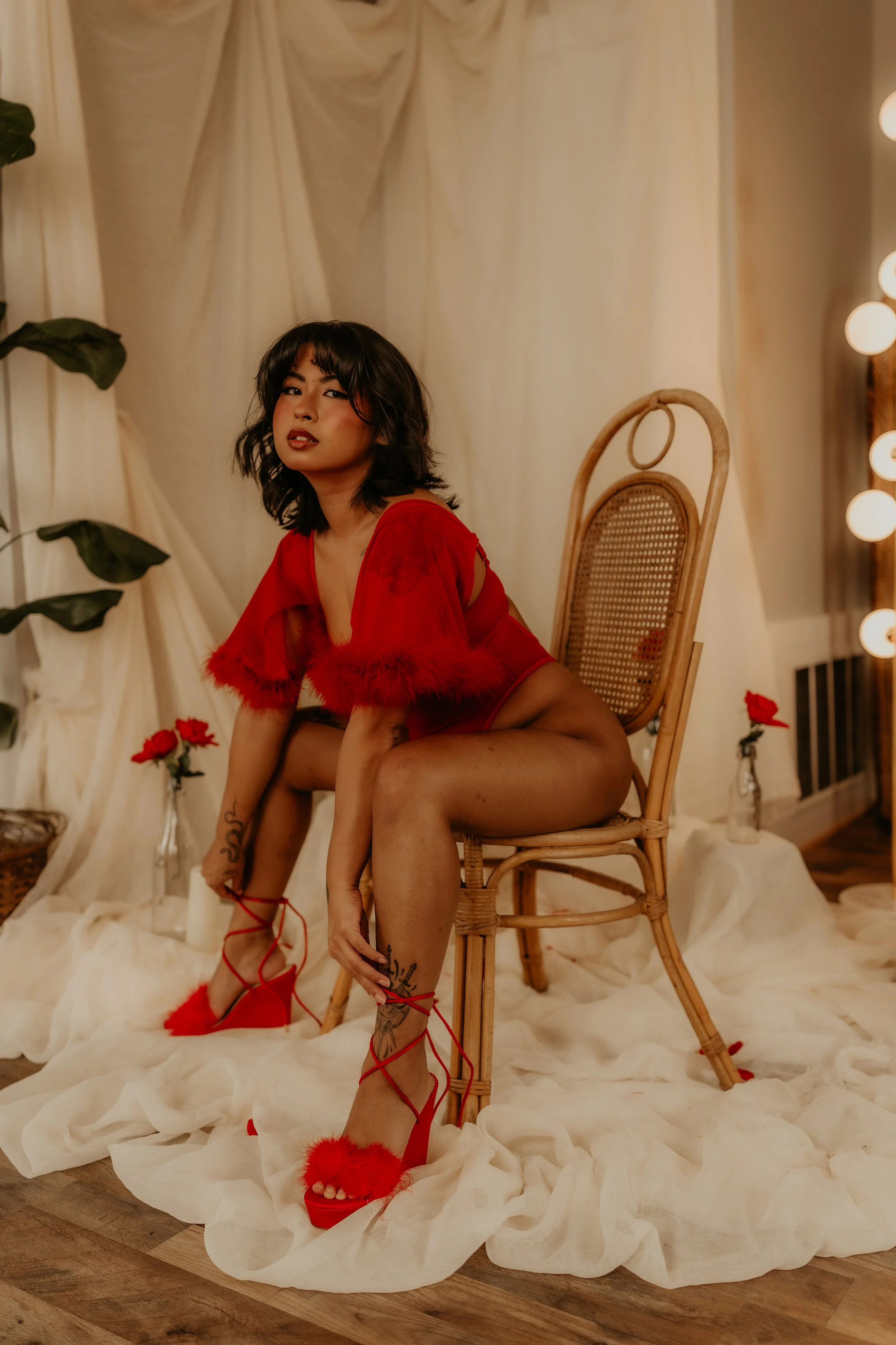 A woman in red lingerie and high heels poses on a wooden chair in a decorated room with cream-colored drapery, red roses, and a soft fabric covering the floor.