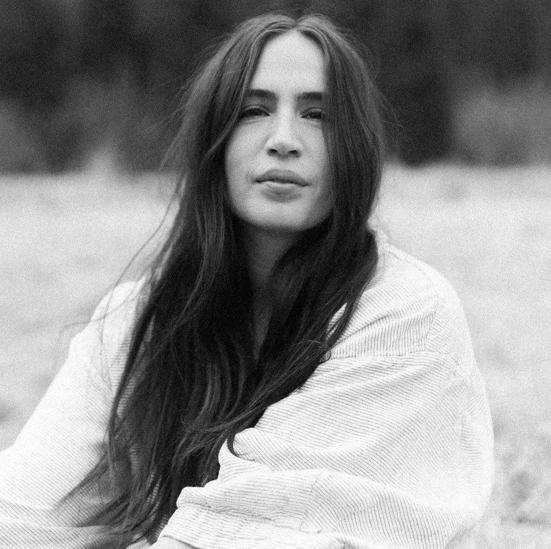 A black and white portrait of a woman with long dark hair, outdoors, with a blurred background.